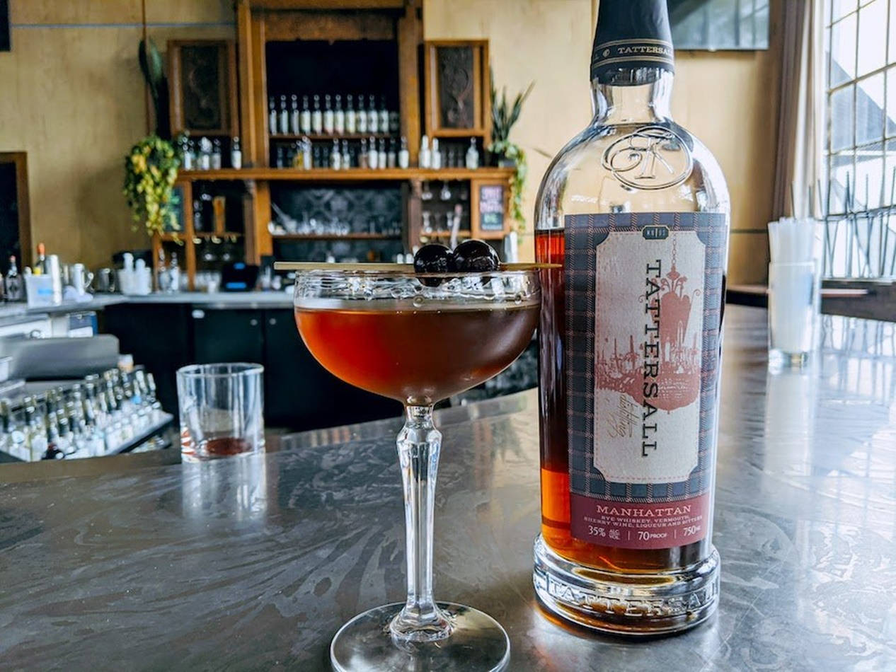 Drink 桌布 壁紙 — 一杯豐富的琥珀色曼哈頓雞尾酒，飾以櫻桃，放在其酒瓶旁邊，在溫暖燈光的酒廊中拋光吧檯上。