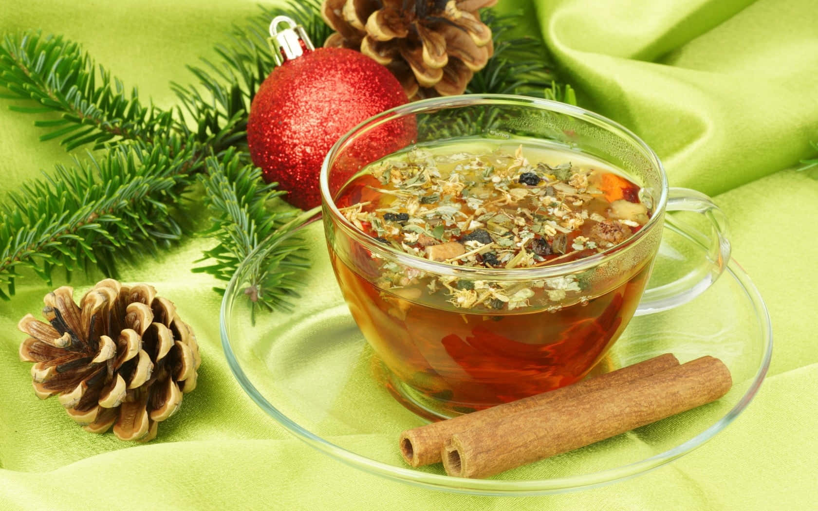 Mulled Wine 桌布 壁紙 — 一杯透明玻璃杯裝的香料草本茶，帶有漂浮的植物成分，放在碟子上，旁邊有肉桂棒、松果和一個紅色裝飾品，置於柔軟的綠色布料上。