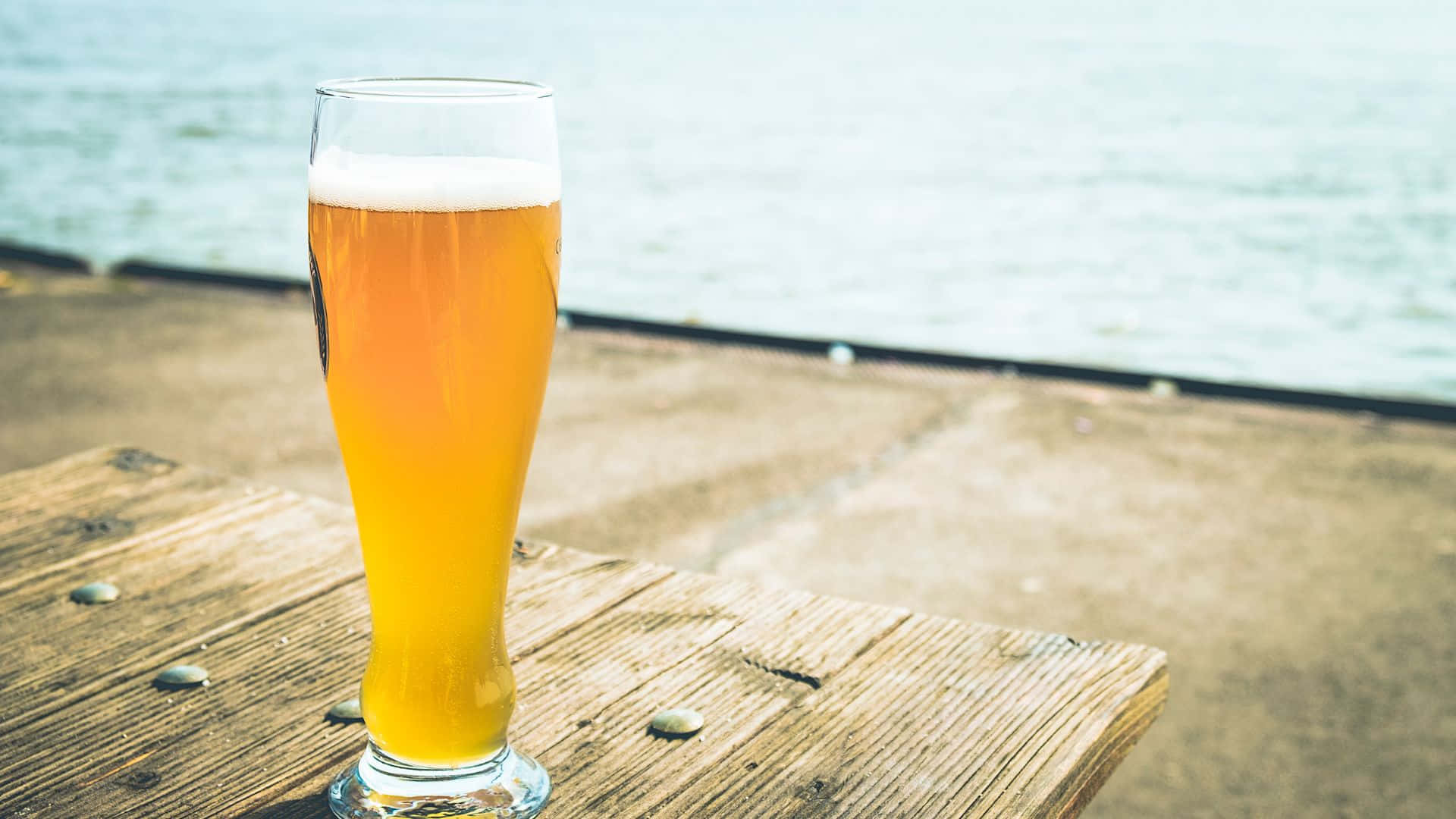 Beer 桌布 壁紙 — 一杯金色啤酒，上面有白色泡沫，放在一張風化的木桌上，旁邊是平靜的水邊。