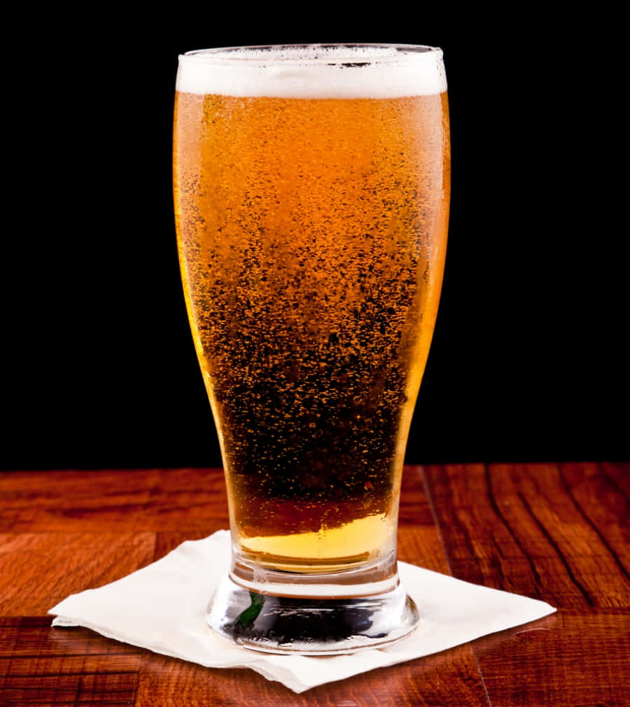Beer 桌布 壁紙 — 一杯金色啤酒，帶有白色泡沫，放在黑色背景的木桌上的一張餐巾上。