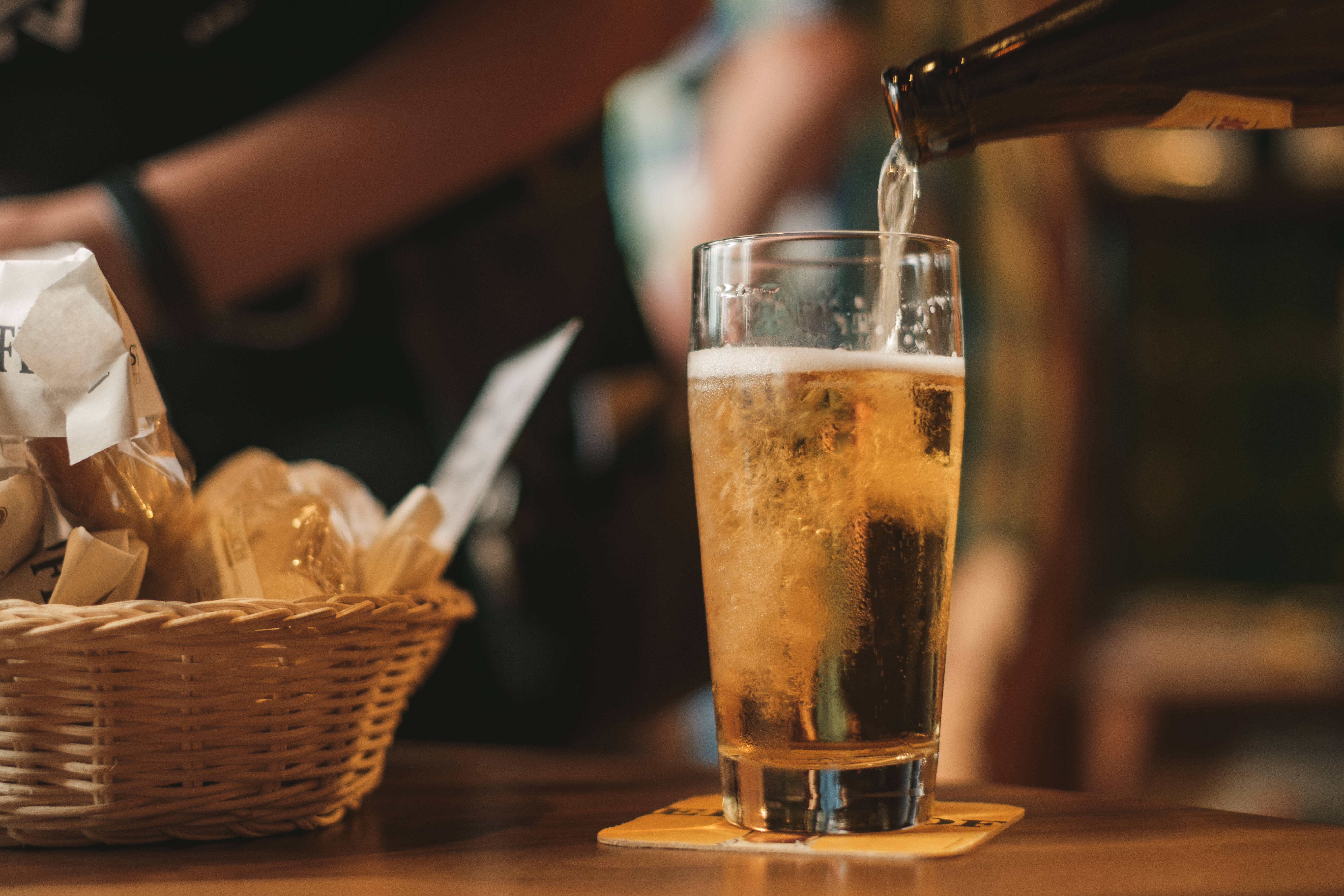 Beer 桌布 壁紙 — 一杯金色啤酒正倒入玻璃杯中，放在木製吧檯上，旁邊有一籃麵包，背景有模糊的酒保。