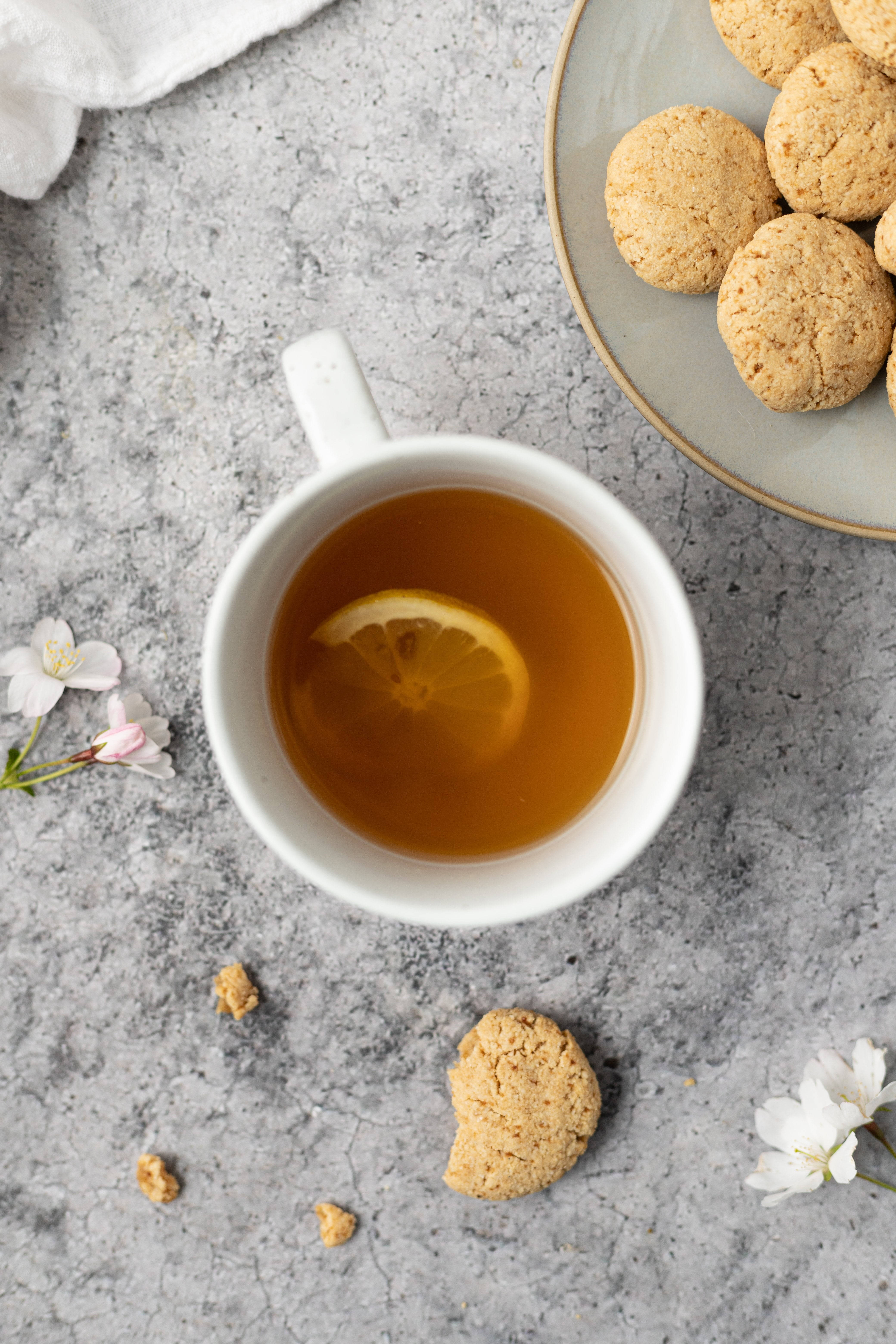 Cookie 桌布 壁紙 — 一杯金色茶的白杯，附有檸檬片，旁邊放著一盤燕麥餅乾和散落的櫻花，在灰色石面上。