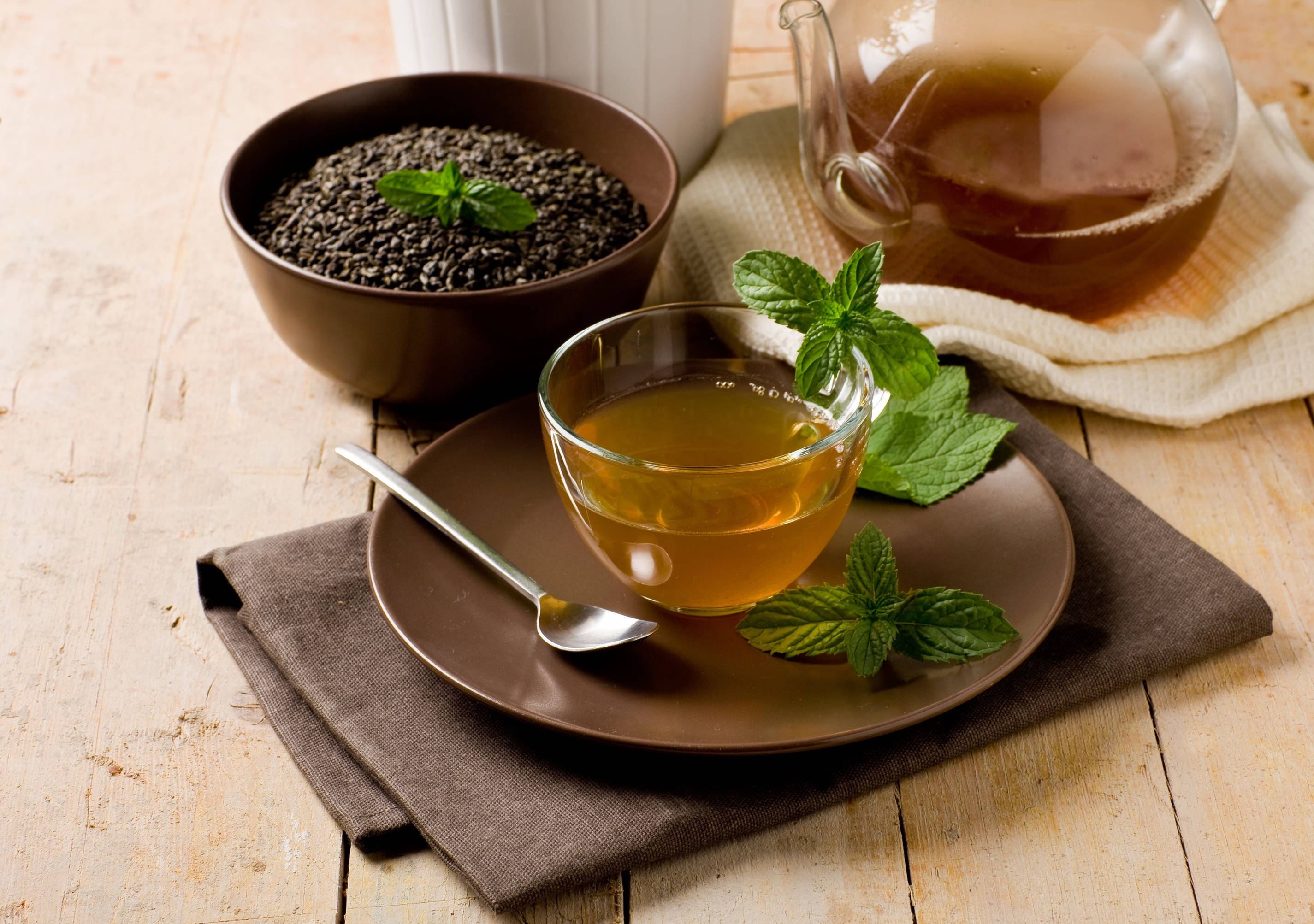 tea 桌布 壁紙 — 一杯金色薄荷茶放在棕色盤子上，旁邊有新鮮薄荷葉和一碗乾薄荷籽，置於木桌上。
