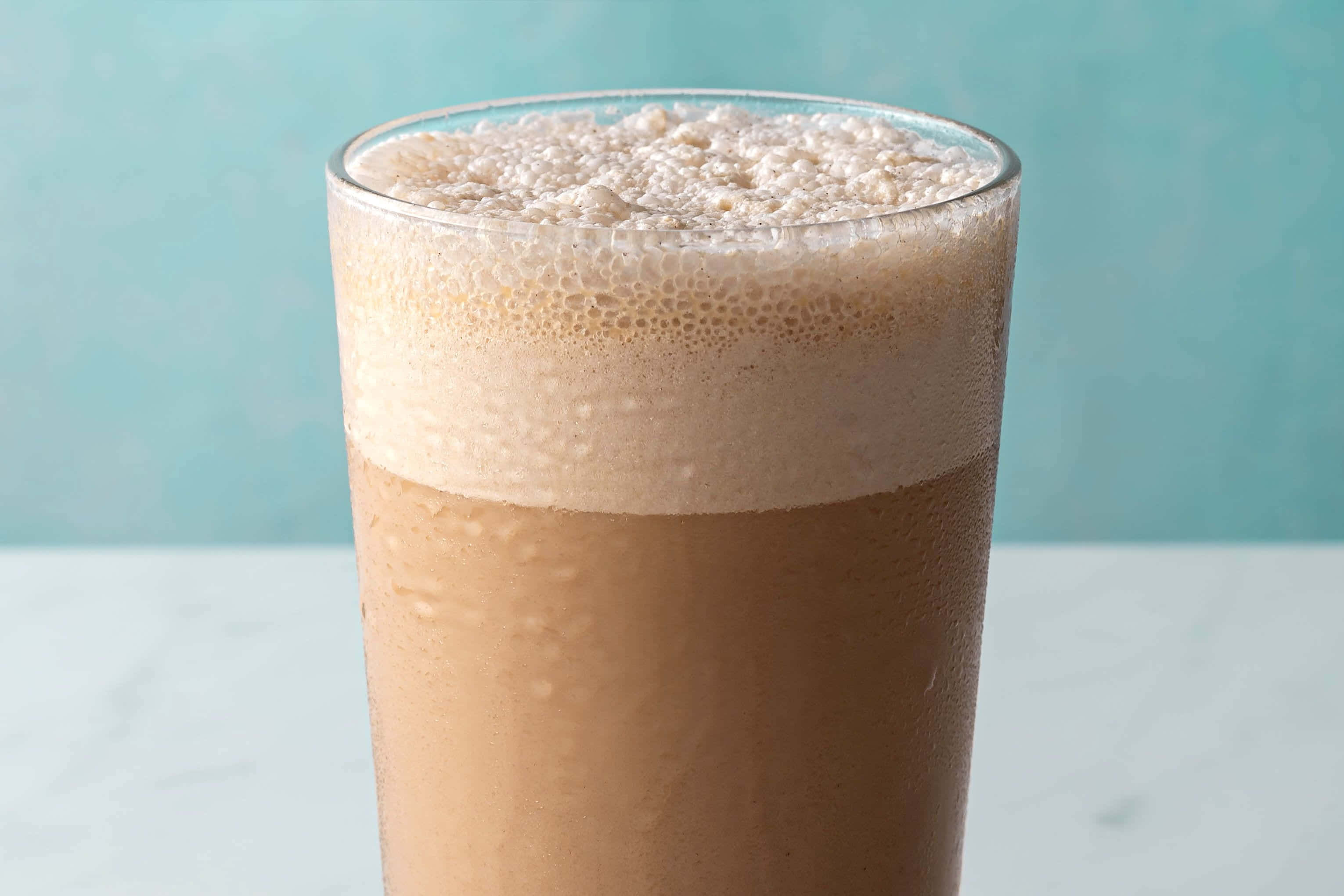 Iced Coffee 桌布 壁紙 — 一杯高身泡沫冰咖啡，帶有水珠，放在白色大理石表面上，背景是柔和的青綠色。