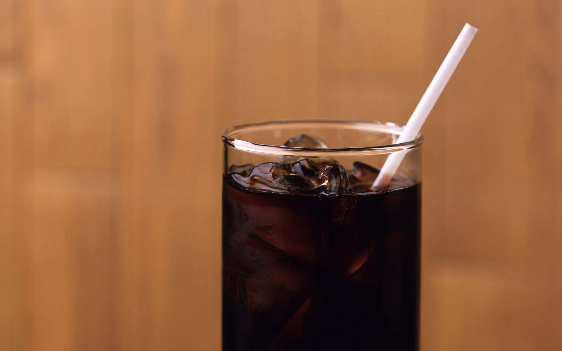 Iced Coffee 桌布 壁紙 — 一杯高身深色冰凍蘇打，有冰塊和白色吸管，襯托溫暖木質背景。