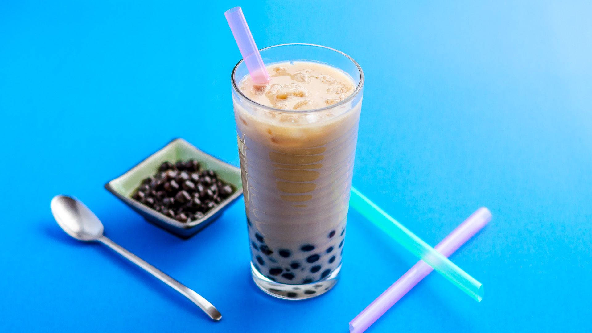 Bubble 桌布 壁紙 — 一杯高身的冰珍珠奶茶，配有木薯珍珠和粉紅色吸管，旁邊放著一小碟珍珠和彩色吸管，背景為明亮的藍色。