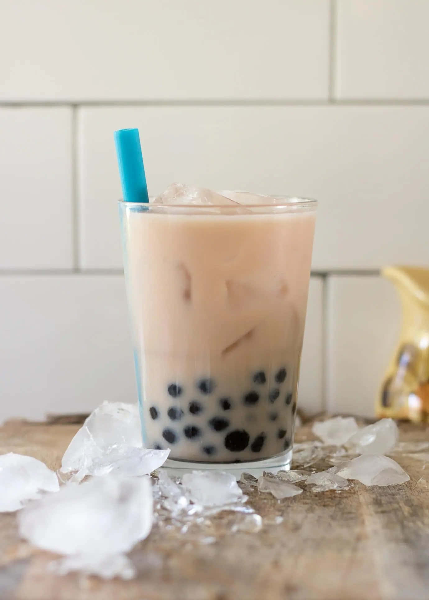 Boba Tea 桌布 壁紙 — 一杯高身的奶油冰珍珠奶茶，有黑色珍珠和藍色吸管，放在木質表面上，周圍有正在融化的冰塊。