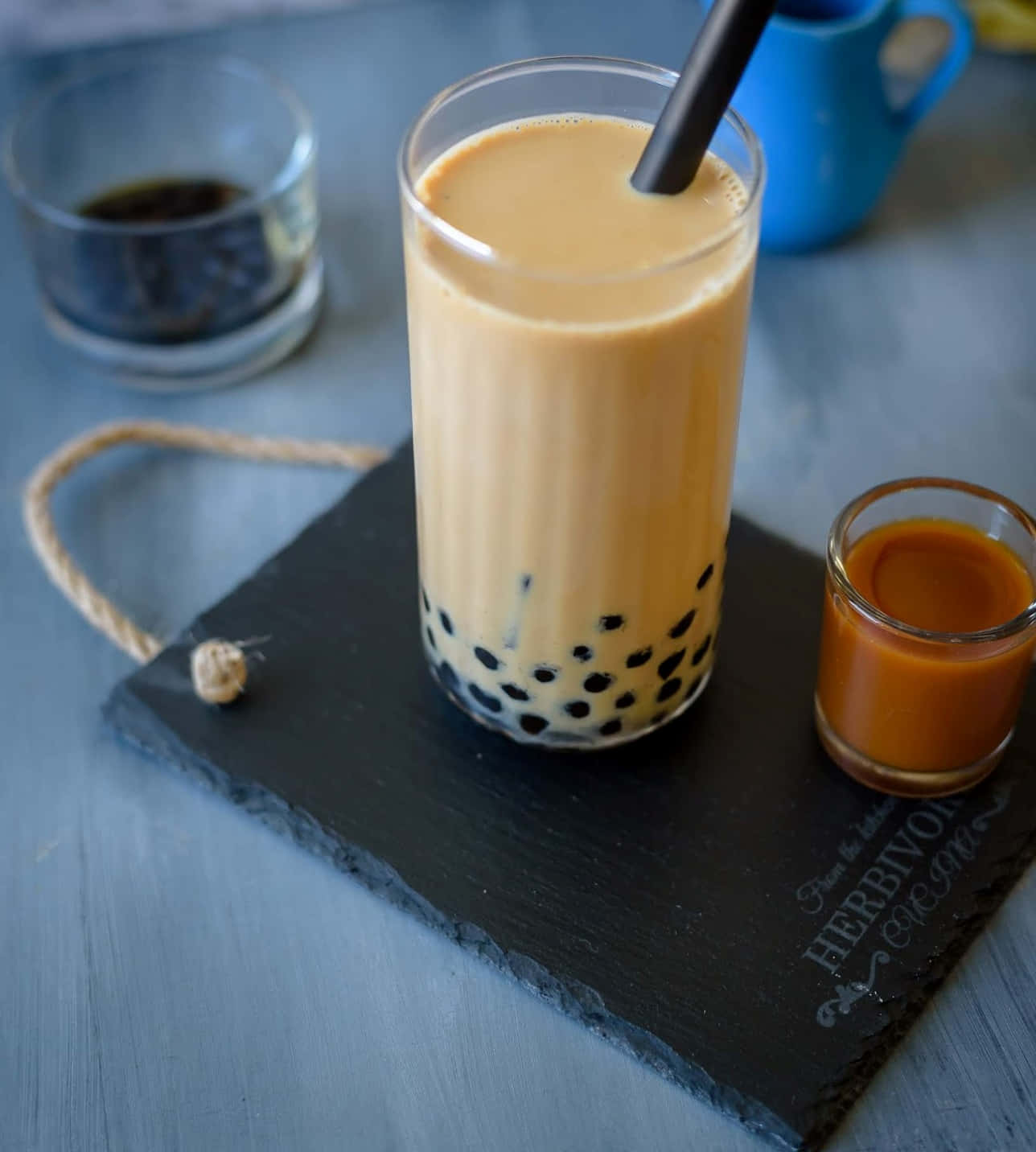 Boba Tea 桌布 壁紙 — 一杯高身的奶油泡泡茶搭配黑色木薯珍珠，放在石板托盤上，旁邊有焦糖醬和藍色馬克杯。