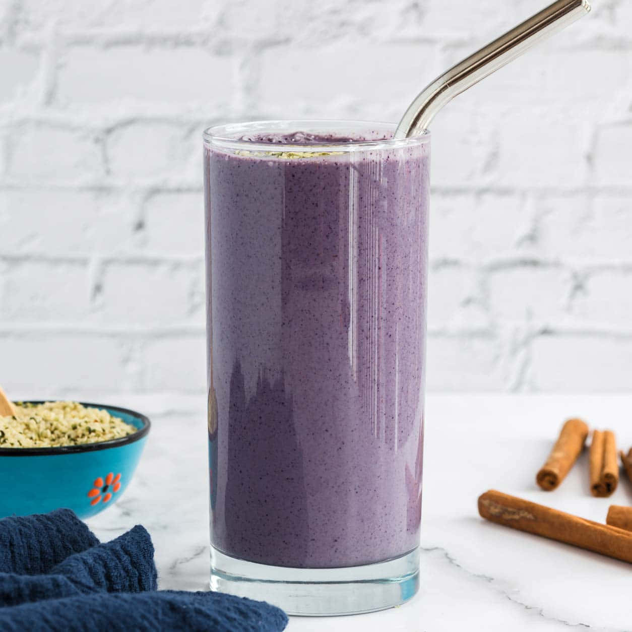 Blueberry Smoothie 桌布 壁紙 — 一杯高身的紫色冰沙，用金屬吸管，放在白色大理石檯面上，旁邊有肉桂棒和一個藍色碗裝著種子。