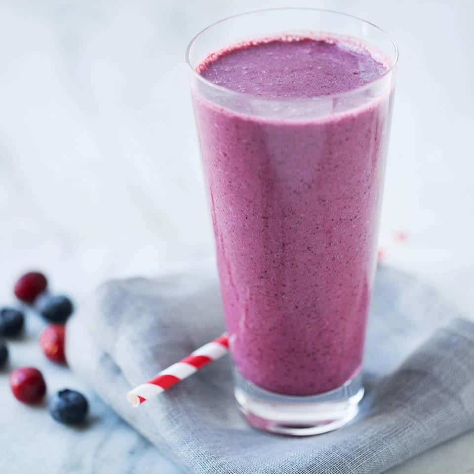 Blueberry Smoothie 桌布 壁紙 — 一杯高身的紫色莓果冰沙，插著紅白條紋吸管，放在灰色布上，旁邊散落著藍莓和蔓越莓。