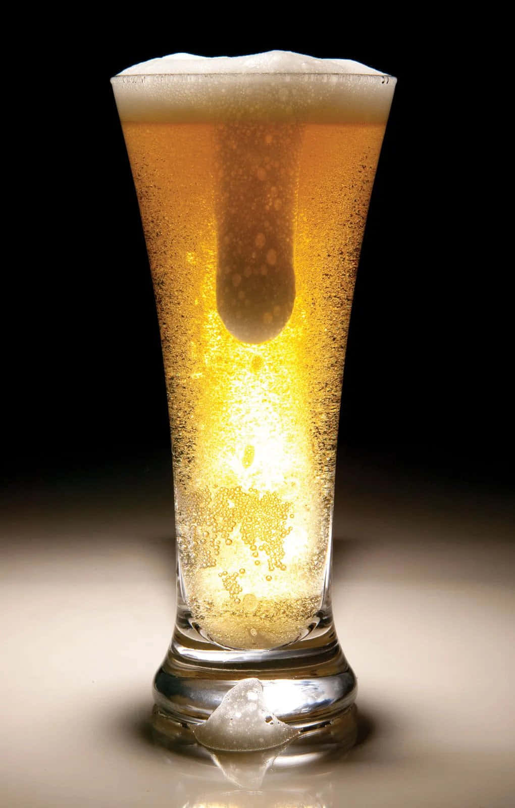 Beer 桌布 壁紙 — 一杯高身金黃色啤酒，帶有泡沫和冰塊，在黑色背景上溫暖地發光。