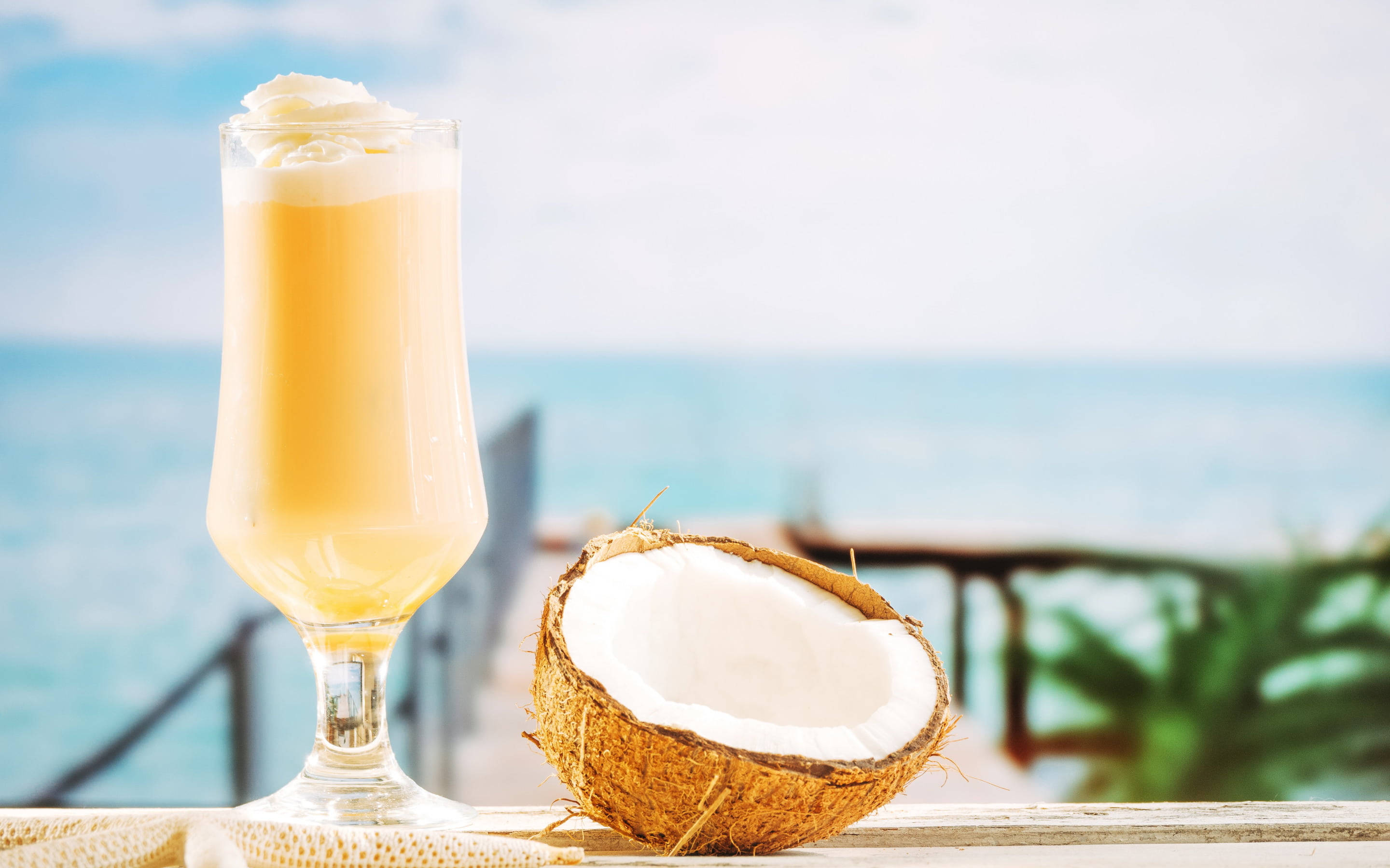 Coconut 桌布 壁紙 — 一杯高高的奶油椰子飲品，上面有鮮奶油，旁邊放著半個椰子，放在木製欄杆上，俯瞰著碧綠色的海洋。