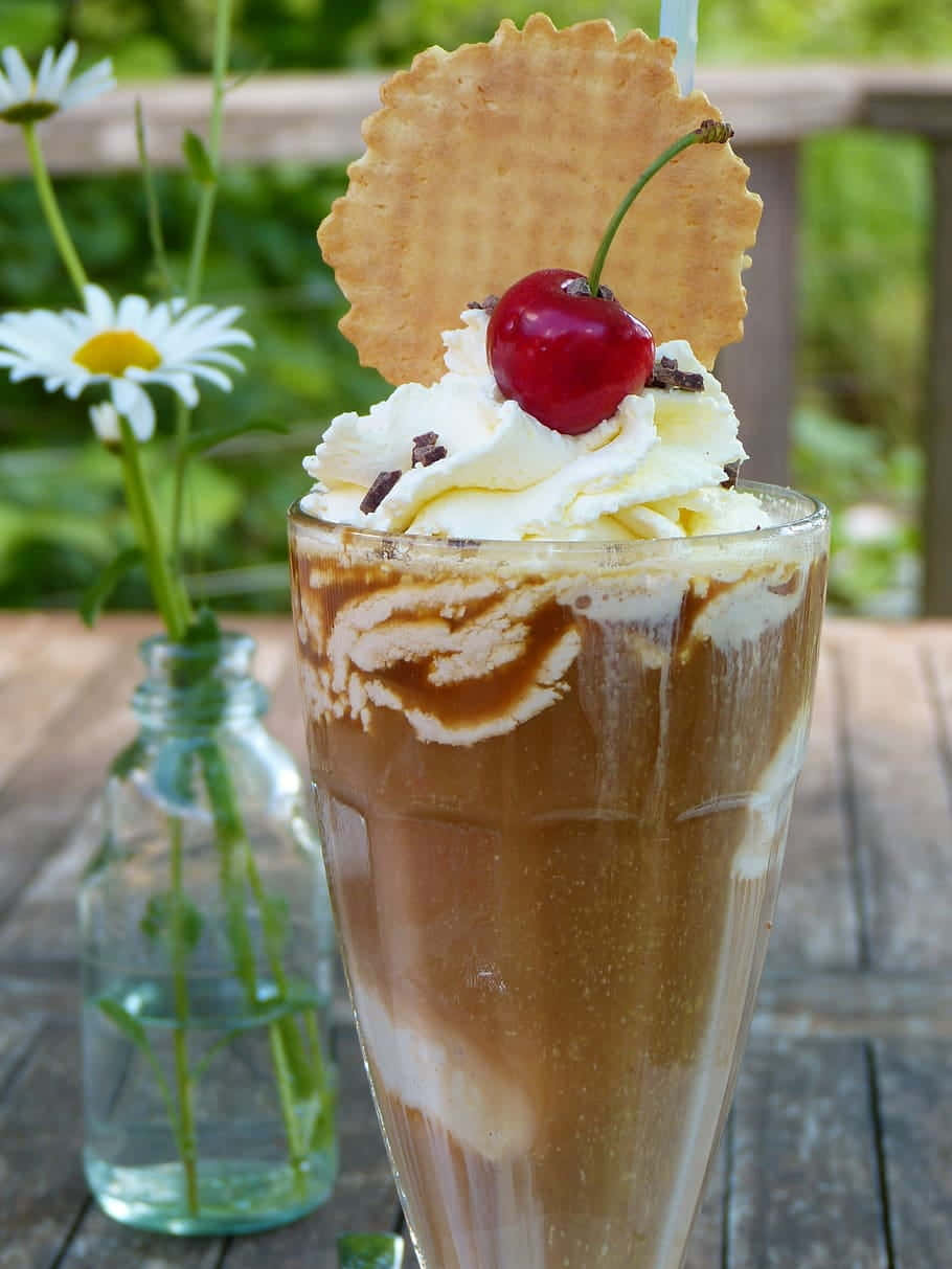 Iced Coffee 桌布 壁紙 — 一杯高高的焦糖漩渦奶昔，上面有鮮奶油、巧克力刨花、一顆櫻桃和一片威化餅，放在木桌上，旁邊有一瓶雛菊。