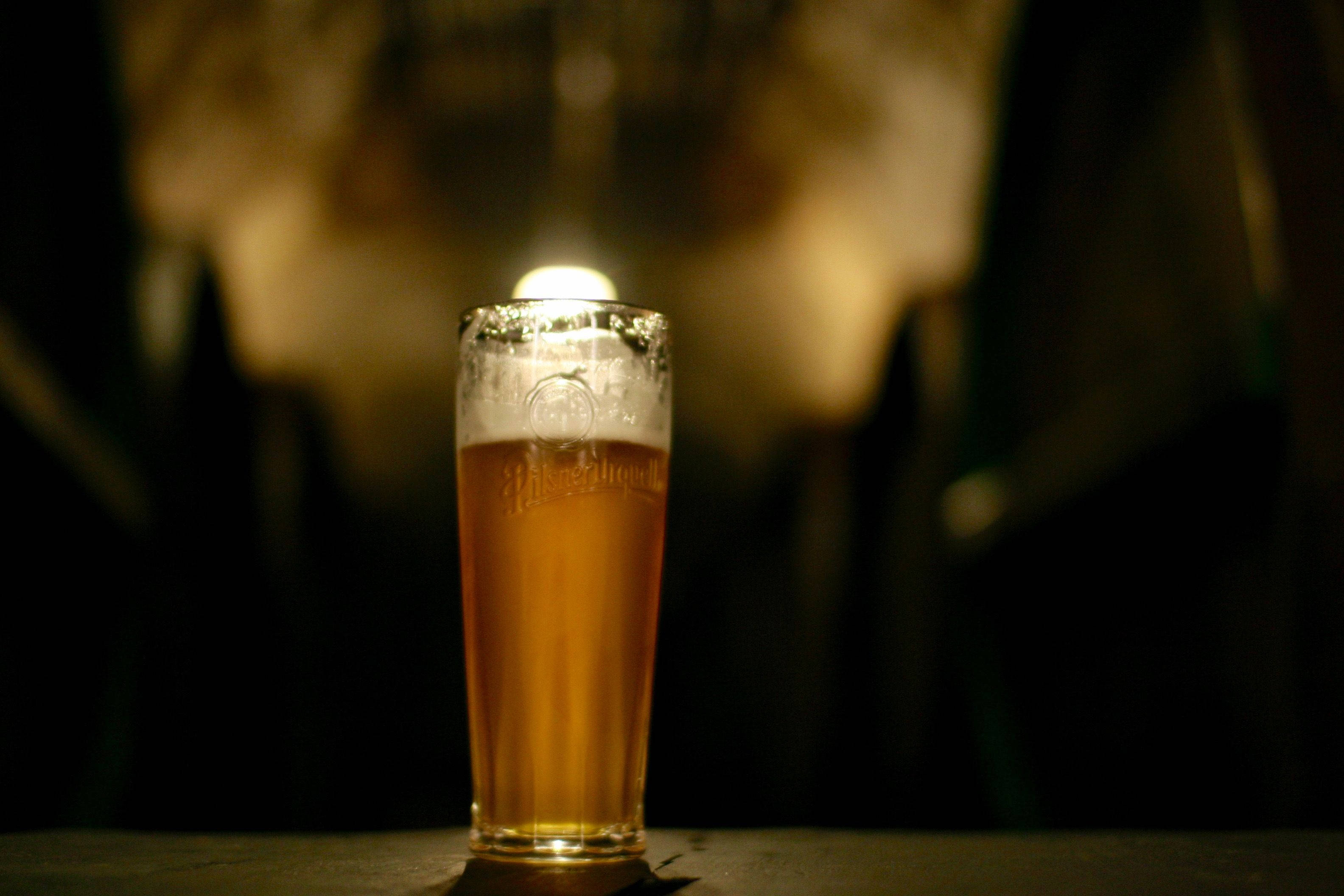 Beer 桌布 壁紙 — 一杯高高的琥珀色啤酒，帶有泡沫，在黑暗的吧檯上，單一聚光燈下溫暖地發光。
