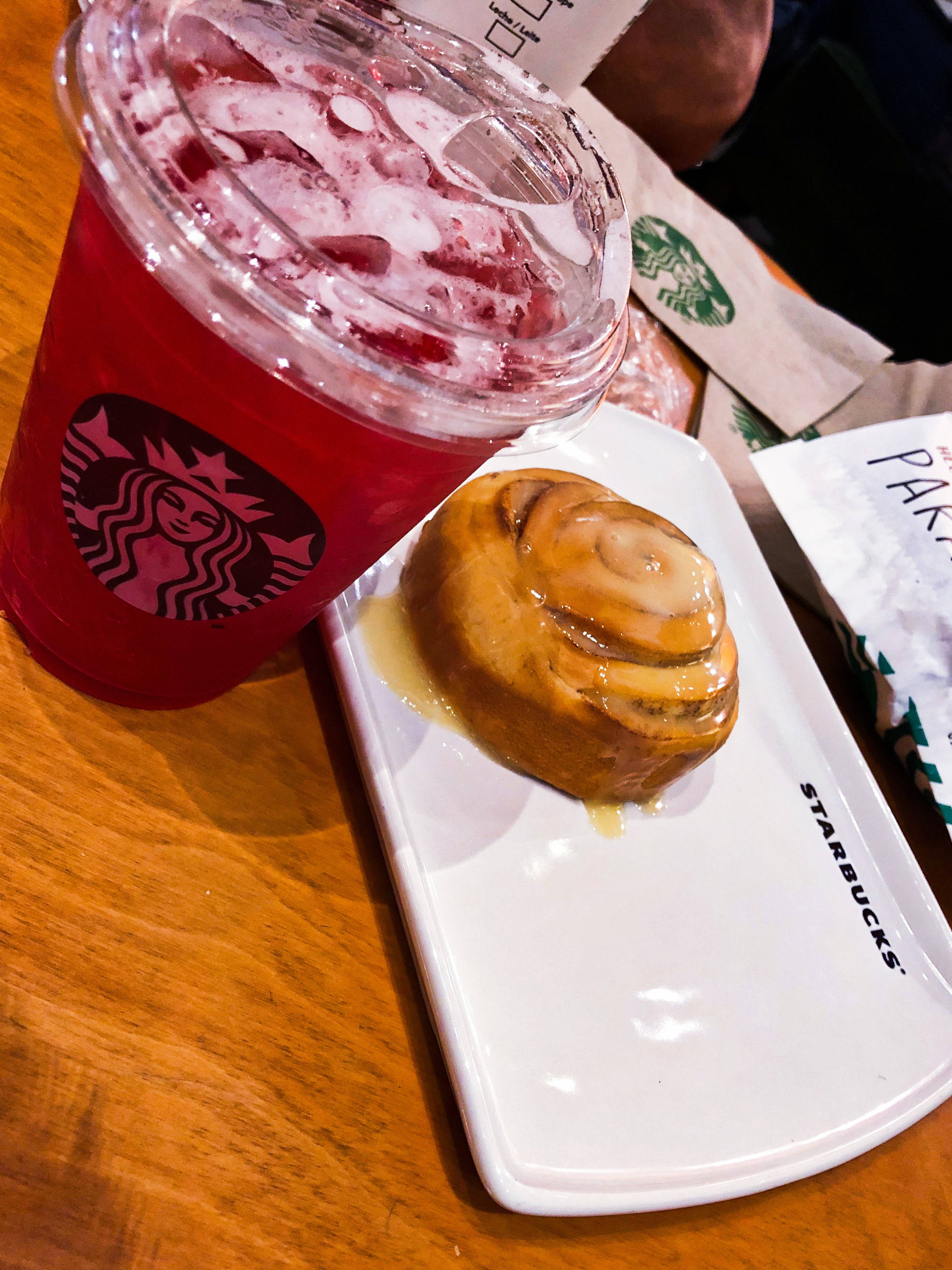 Pastry 桌布 壁紙 — 一杯鮮紅色的冰凍飲品和一個黏稠的肉桂卷放在溫暖木桌上的白色Starbucks碟子上。