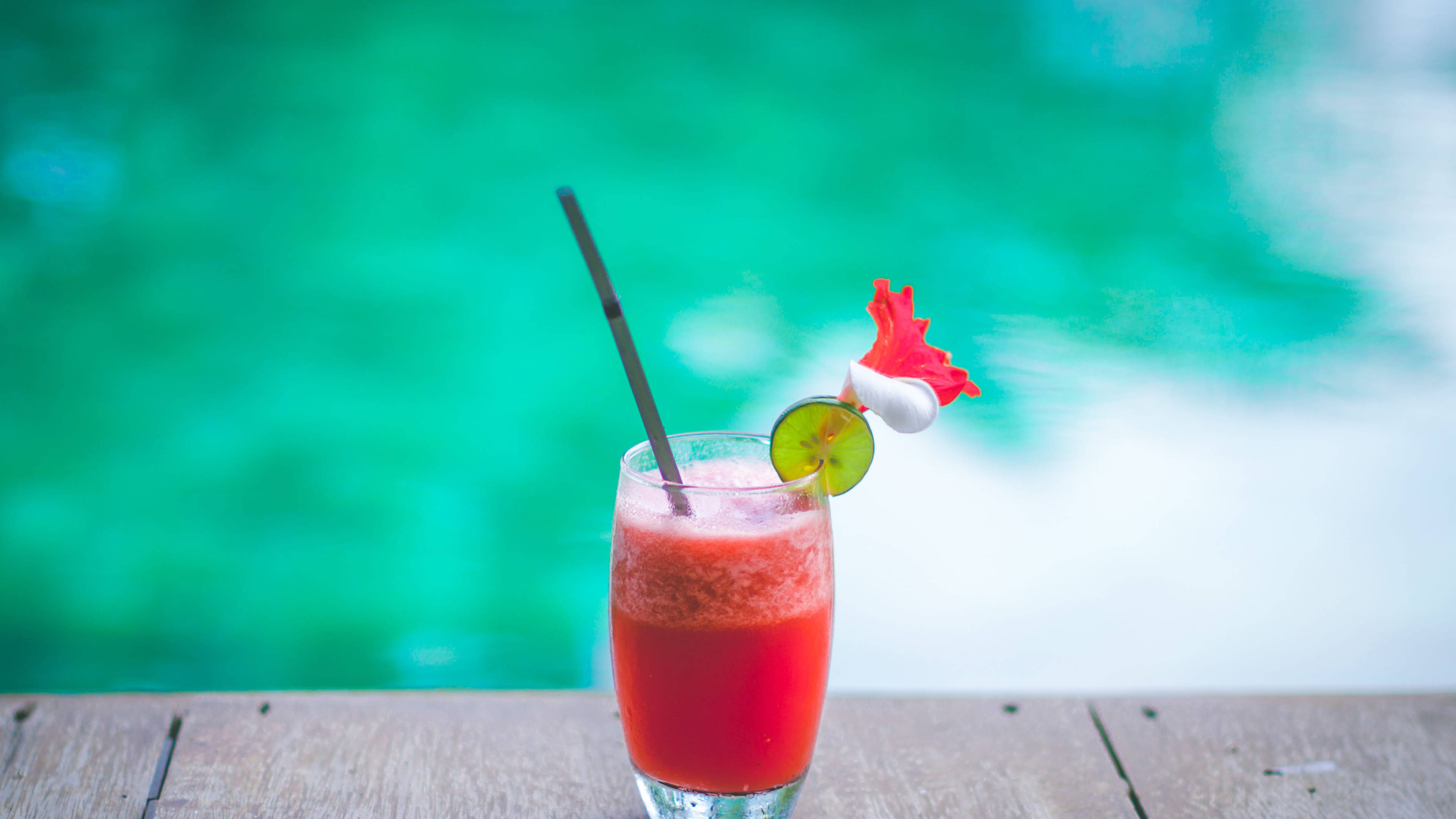 Tropical Drink 桌布 壁紙 — 一杯鮮豔的紅色熱帶飲品，配上萊姆和花朵裝飾，放在木桌上，旁邊是藍綠色的游泳池。