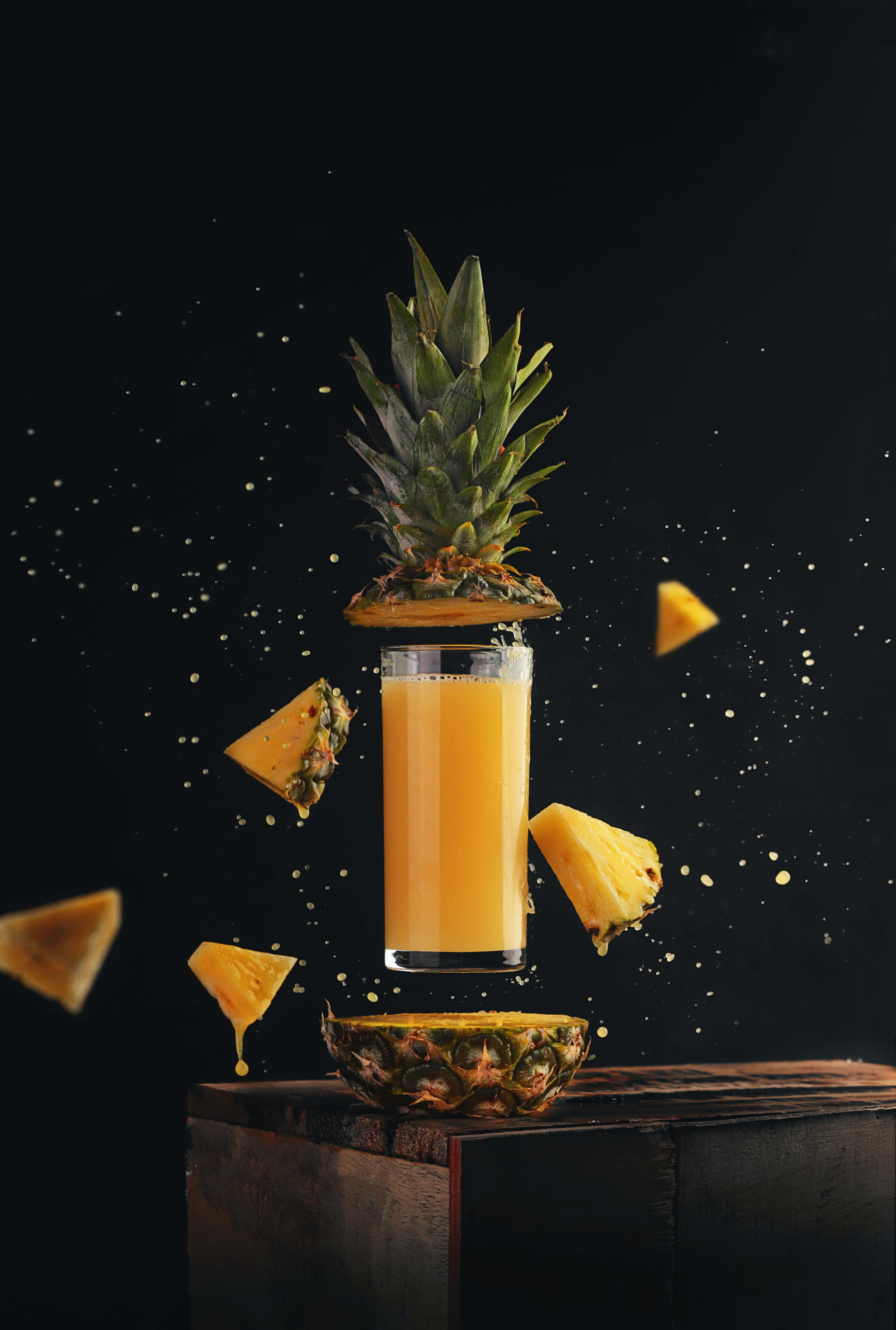 Pineapple 桌布 壁紙 — 一杯鳳梨汁懸浮在半空中，伴隨著漂浮的鳳梨塊和皇冠，背景為黑色。