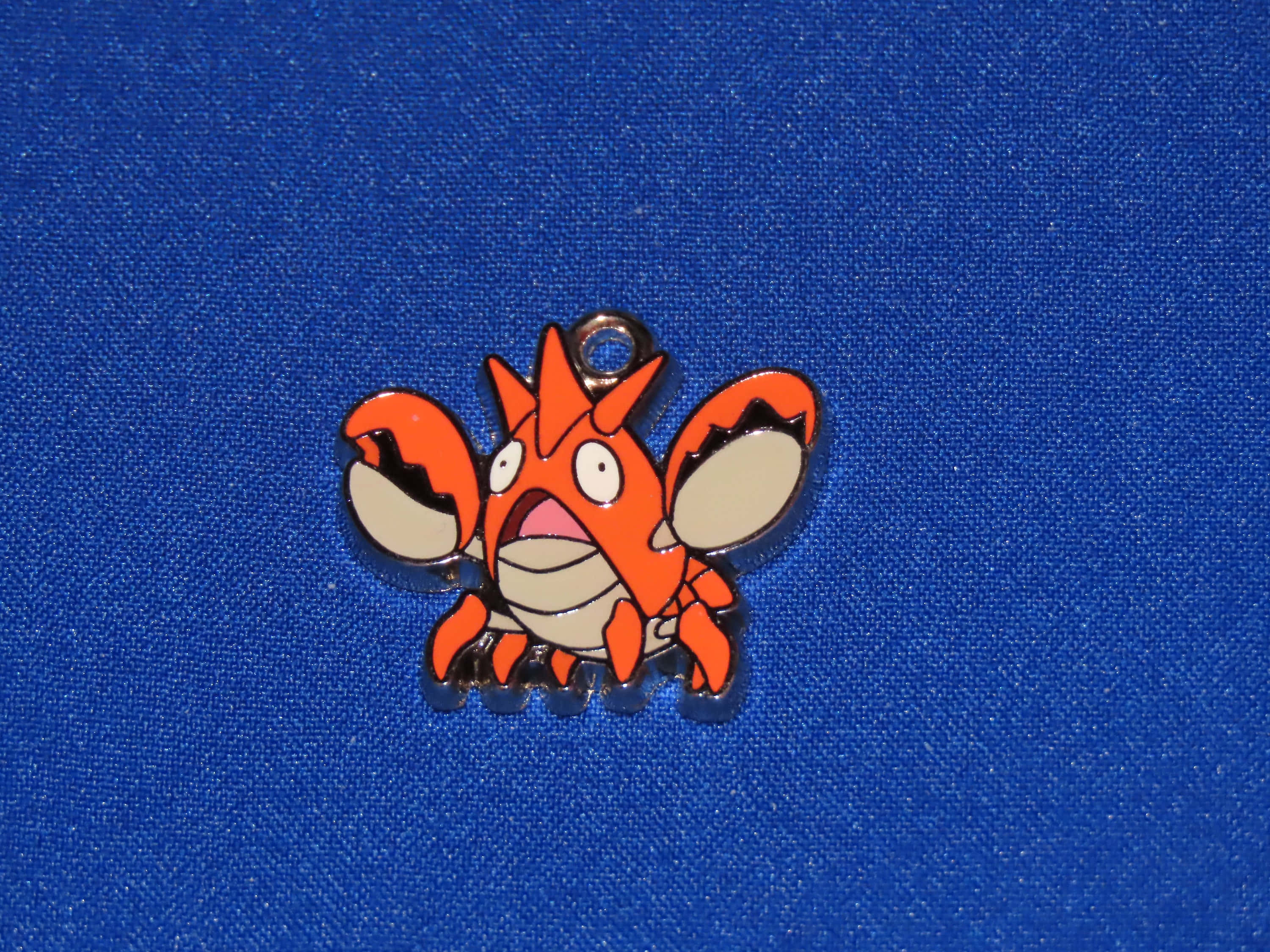 寶可夢 桌布 壁紙 — 一枚亮橙色的Krabby Pokémon琺瑯別針，有白色眼睛和米色爪子，放在有紋理的皇家藍色布料背景上。
