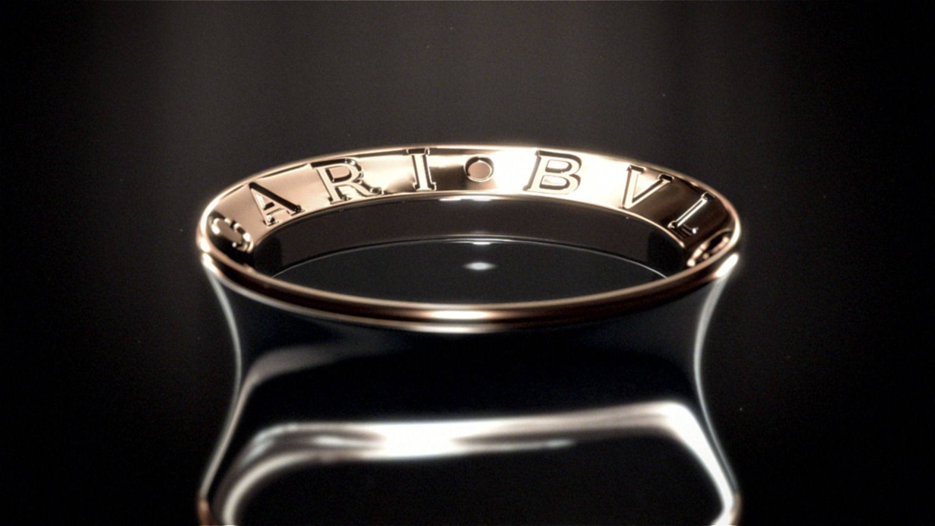 Bvlgari 桌布 壁紙 — 一枚光滑的金戒指，上面刻有字母和一個點，放置在戲劇性燈光下的反光黑色表面上。
