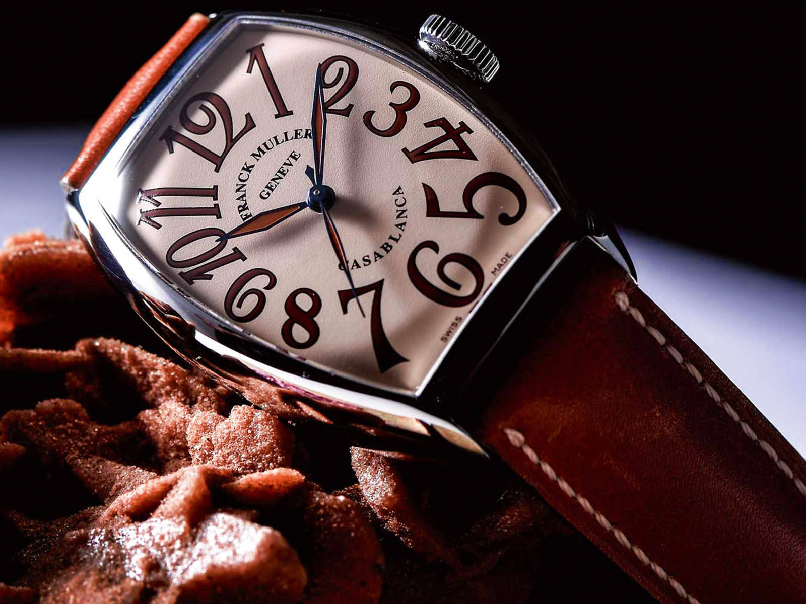 Woman 桌布 壁紙 — 一枚奢華的 Franck Muller 手錶，配備弧形銀色錶殼和棕色皮革錶帶，在戲劇性燈光下優雅地放置在質感豐富的巧克力片上。