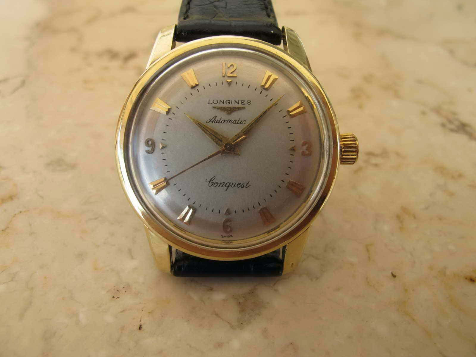 Clock 桌布 壁紙 — 一枚復古金色Longines Conquest自動腕錶搭配黑色皮革錶帶，放置在米色大理石表面上。
