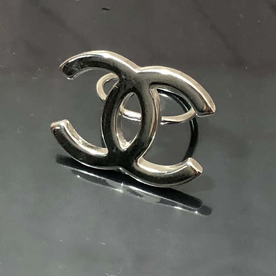 香奈兒標誌 桌布 壁紙 — 一枚時尚的銀色 Chanel 標誌戒指在深色反光表面上閃耀，帶有銳利的高光和柔和的陰影。