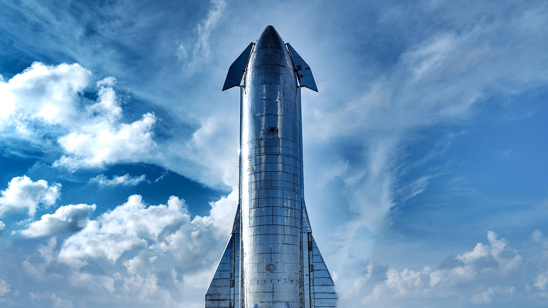Starship 桌布 壁紙 — 一枚流線型的銀色SpaceX Starship火箭，在充滿 fluffy 白色雲朵的鮮豔藍天下高高聳立。