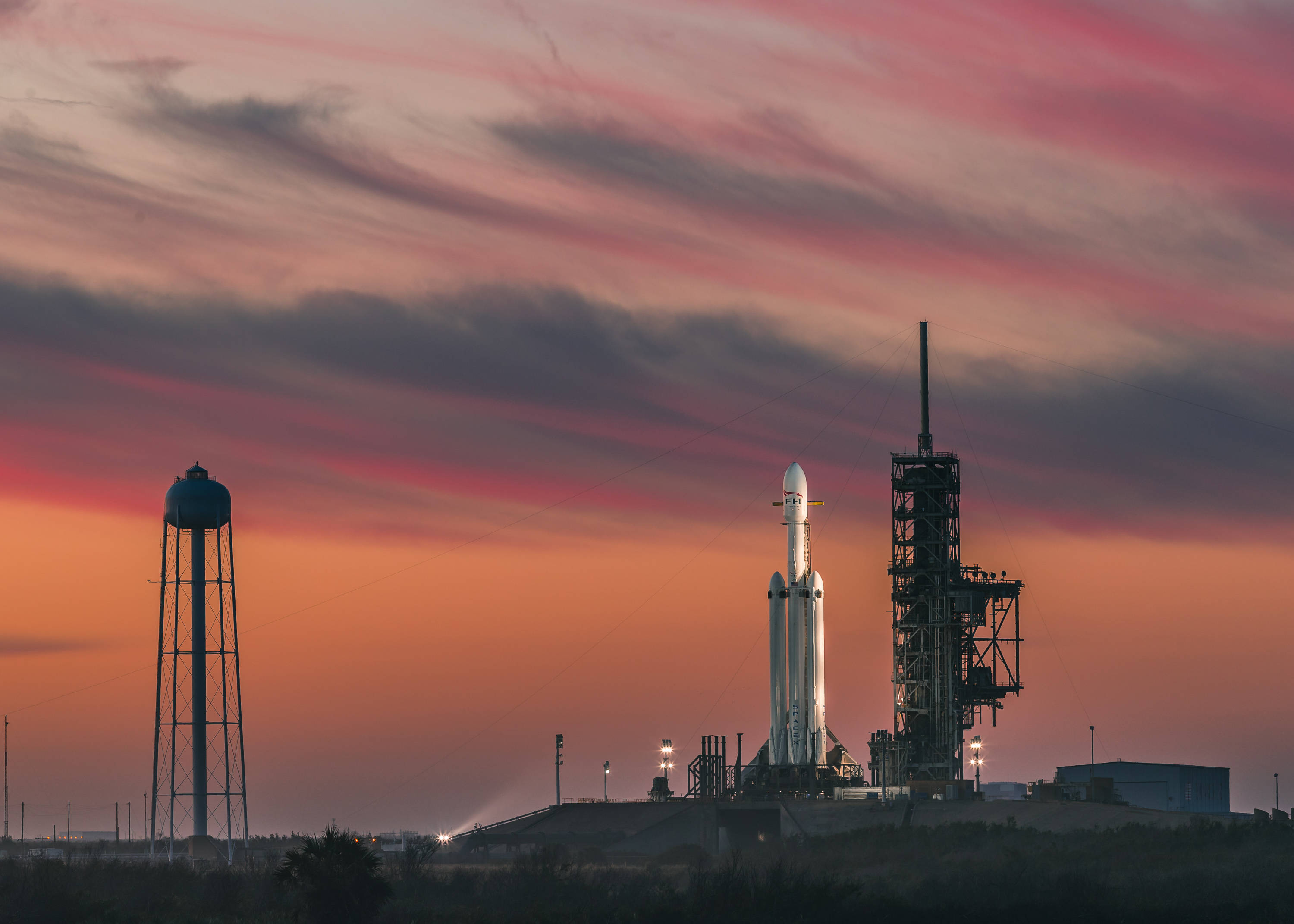 NASA 動態圖片 桌布 壁紙 — 一枚火箭在戲劇性的粉紅色和橙色日落天空下矗立在發射台上，附近有一個水塔。