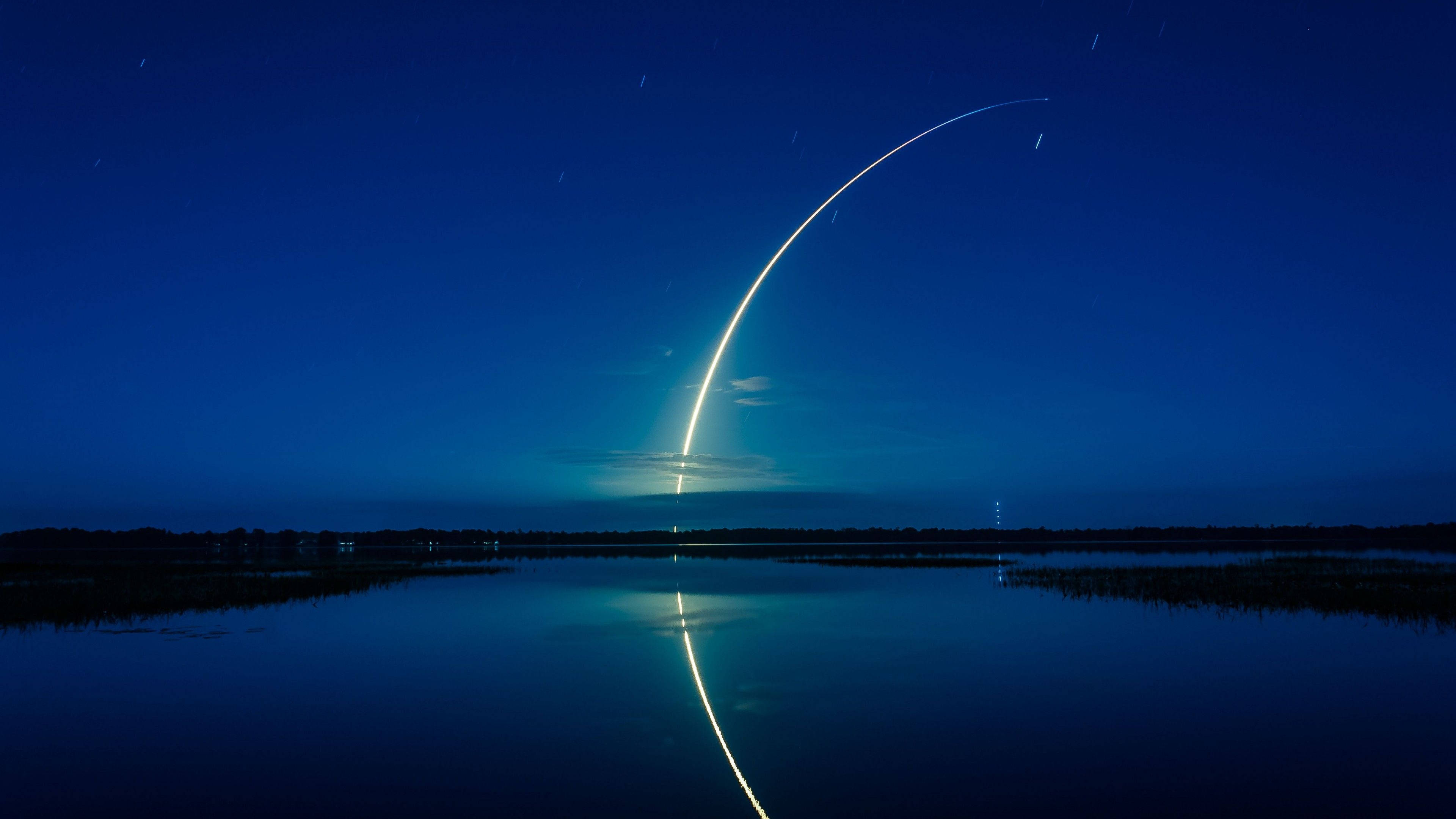 SpaceX 桌布 壁紙 — 一枚火箭在繁星點點的夜空中以燦爛的弧線上升，其倒影完美地閃爍在下方寧靜的湖面上。