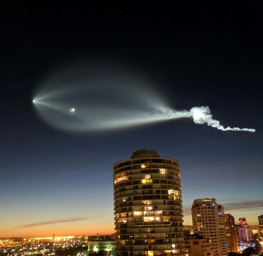 夜空 桌布 壁紙 — 一枚火箭在黃昏時分發射，背景是燈火通明的摩天大樓和暮色天空中發光的尾跡。