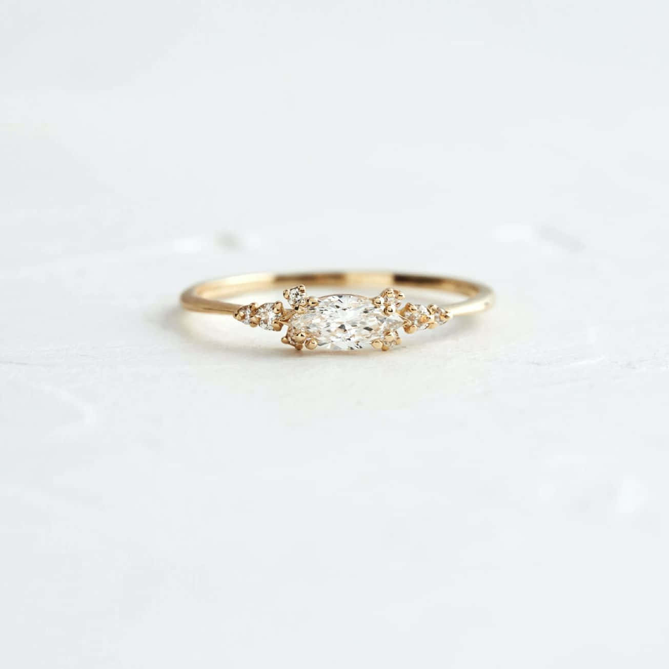 Wedding Rings 桌布 壁紙 — 一枚精緻的金戒指，中央鑲有欖尖形鑽石，周圍鑲有較小的鑽石，置於柔軟的白色紋理表面上。