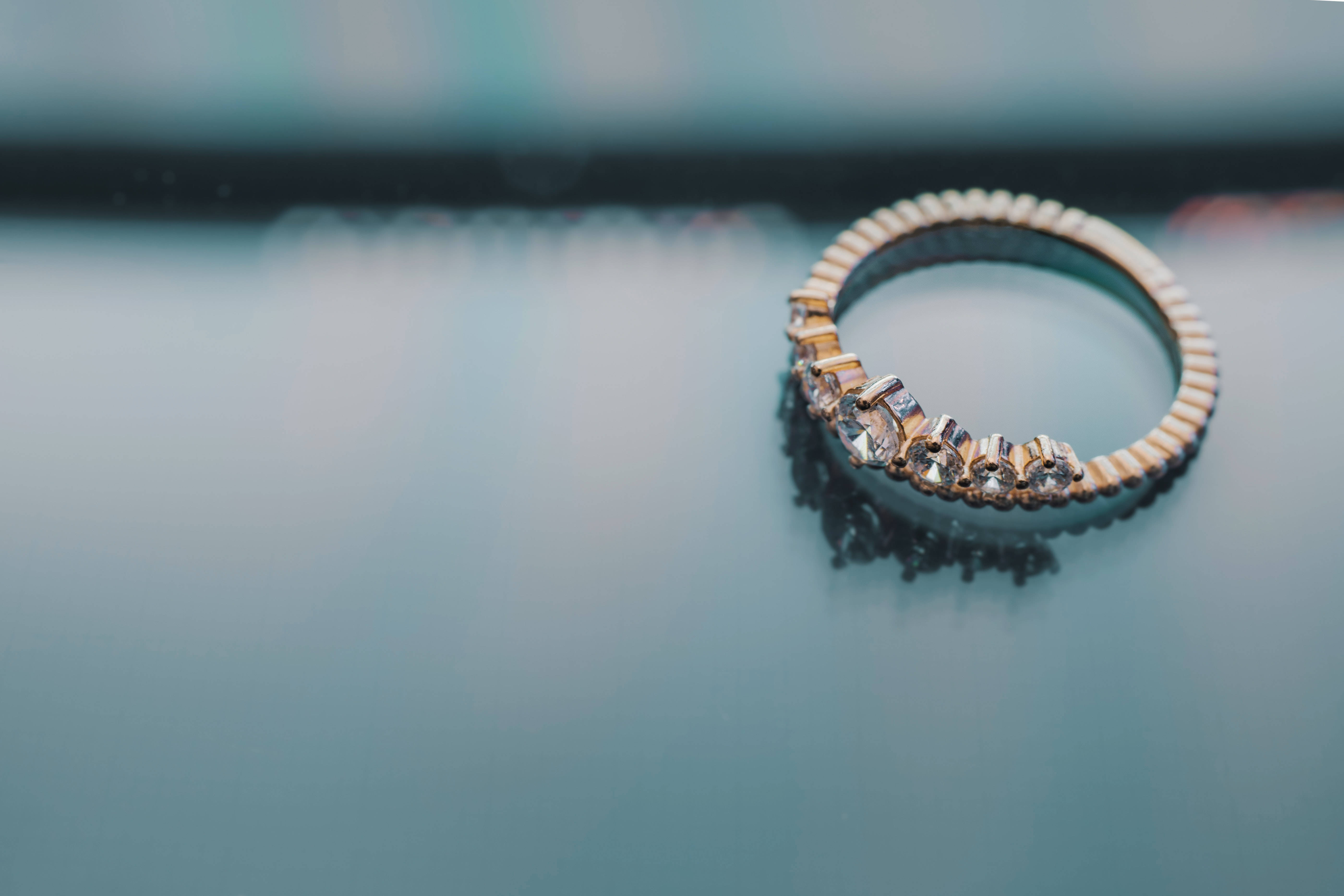 Gold Jewellery 桌布 壁紙 — 一枚精緻的金戒指，鑲有閃爍寶石，放置在清涼的藍色反光表面上，背景柔和模糊。