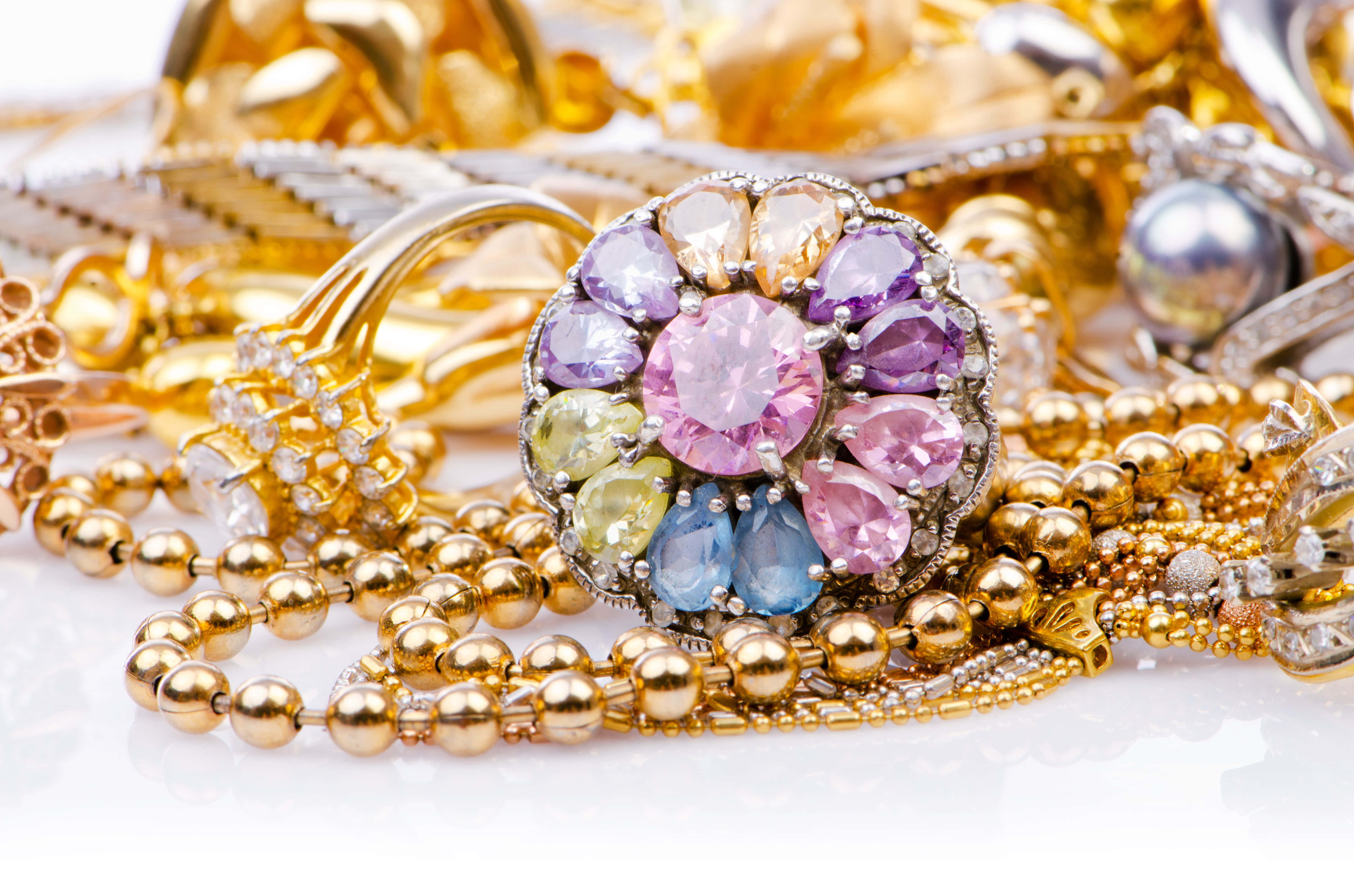 Jewelry Design 桌布 壁紙 — 一枚色彩繽紛的花卉胸針，鑲有粉紅、紫、藍和黃色寶石，放在白色表面上的一堆閃閃發光的黃金首飾之上。