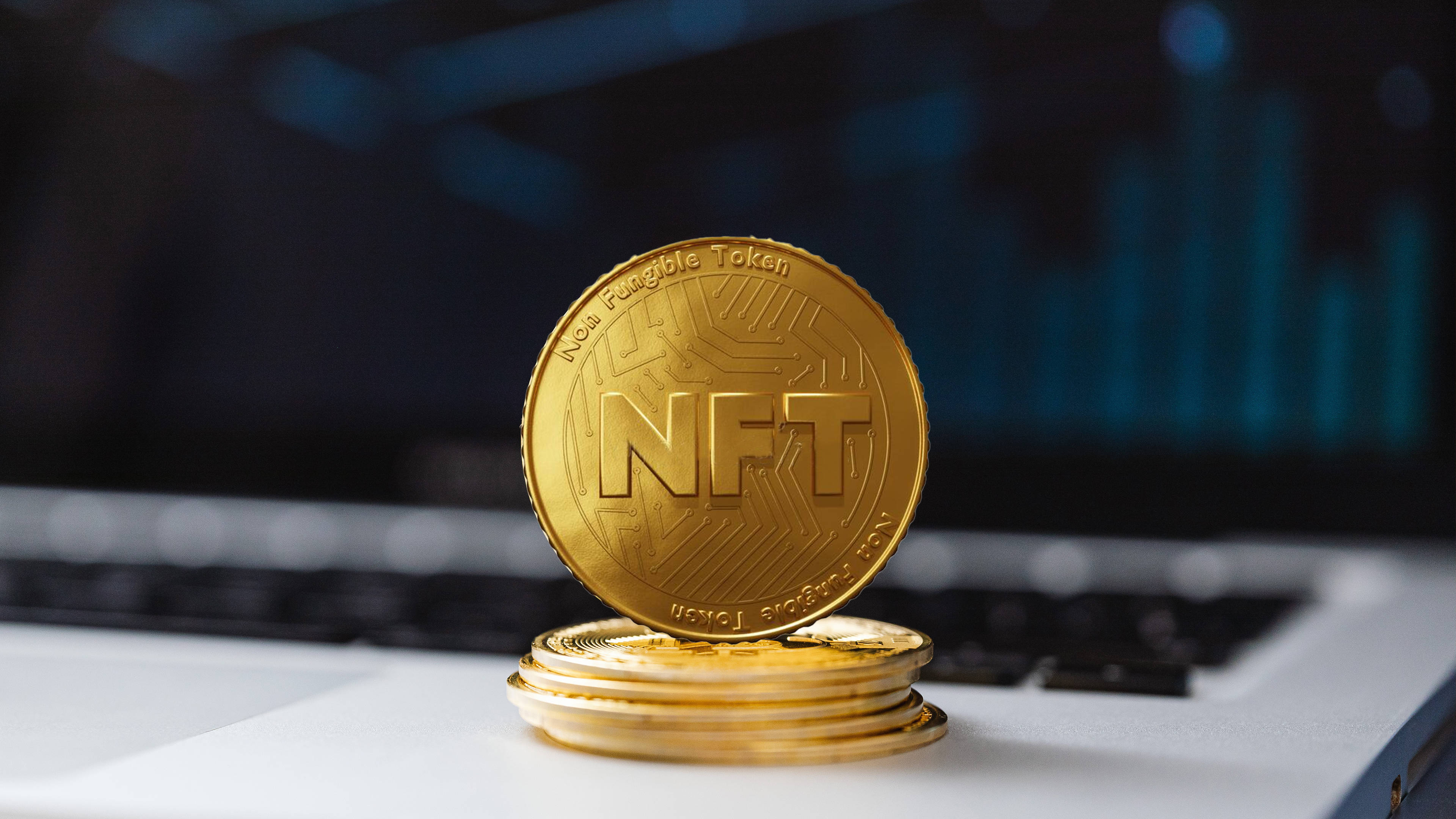 NFT 桌布 壁紙 — 一枚金色NFT硬幣立於筆記型電腦鍵盤上的一疊物品之上，背景是模糊的藍色金融圖表。