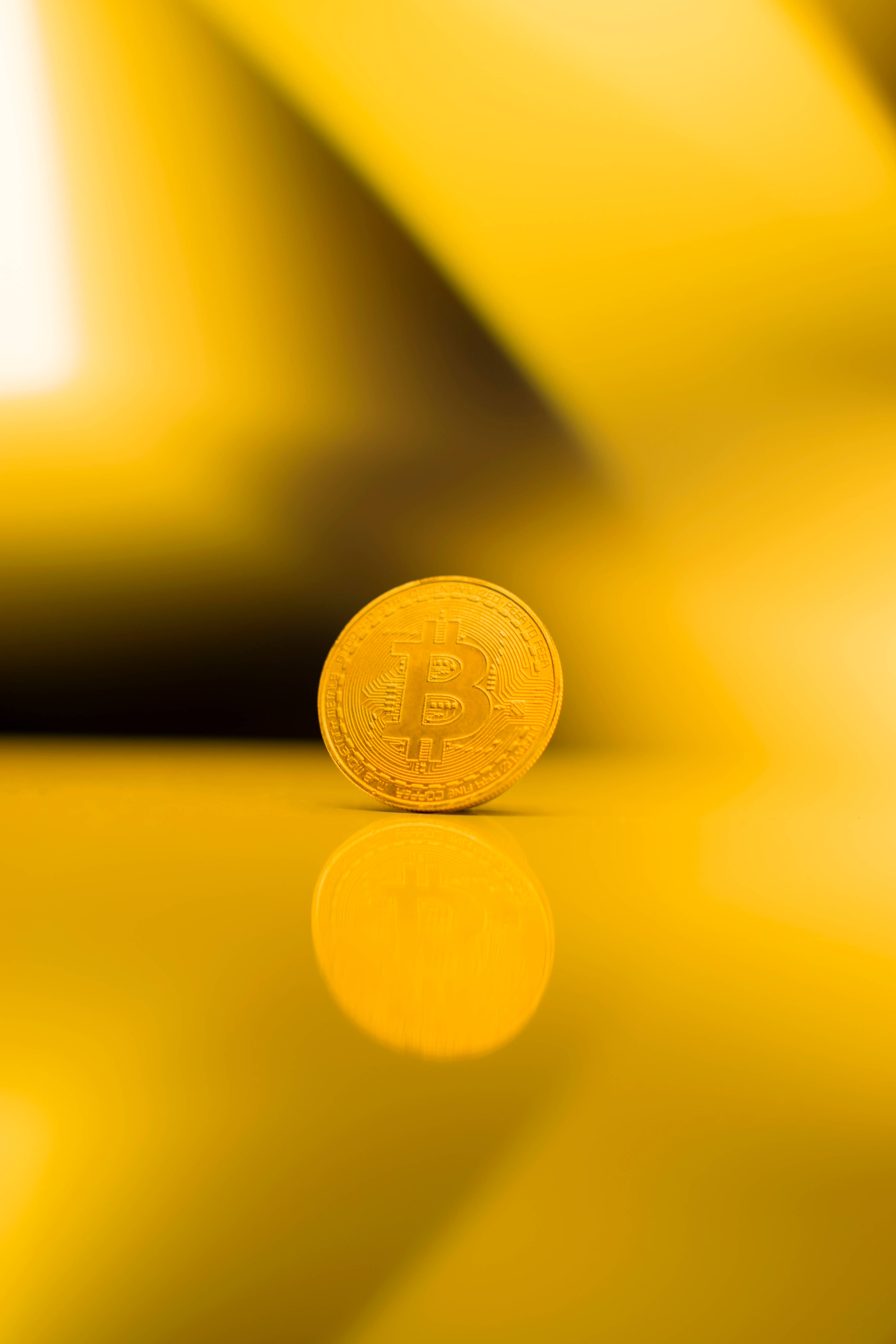 比特幣 桌布 壁紙 — 一枚金色比特幣硬幣放在反射表面上，背景為暖黃色漸變。