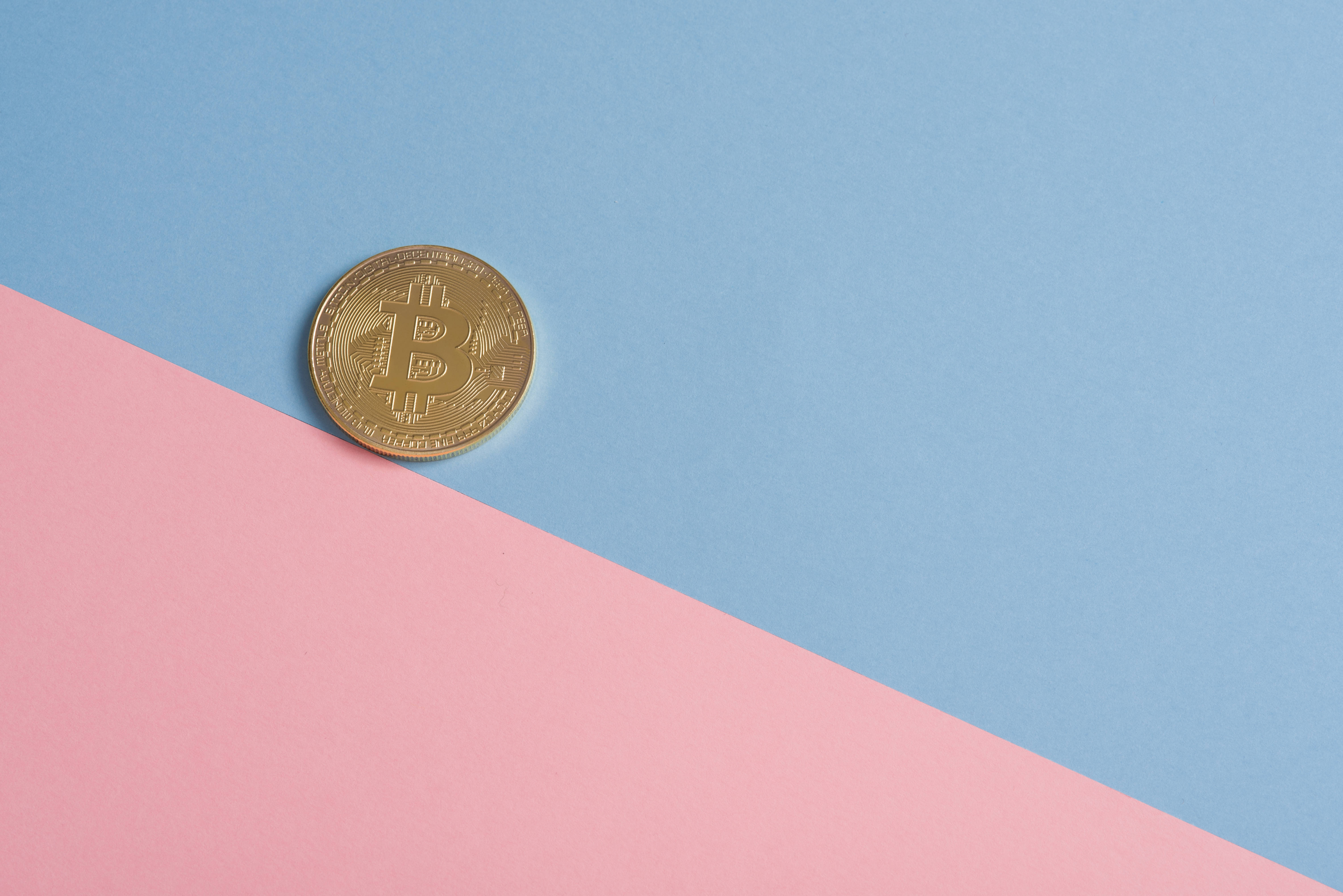 金錢 桌布 壁紙 — 一枚金色比特幣硬幣放在柔軟粉紅色和藍色紙張的對角分割線上。