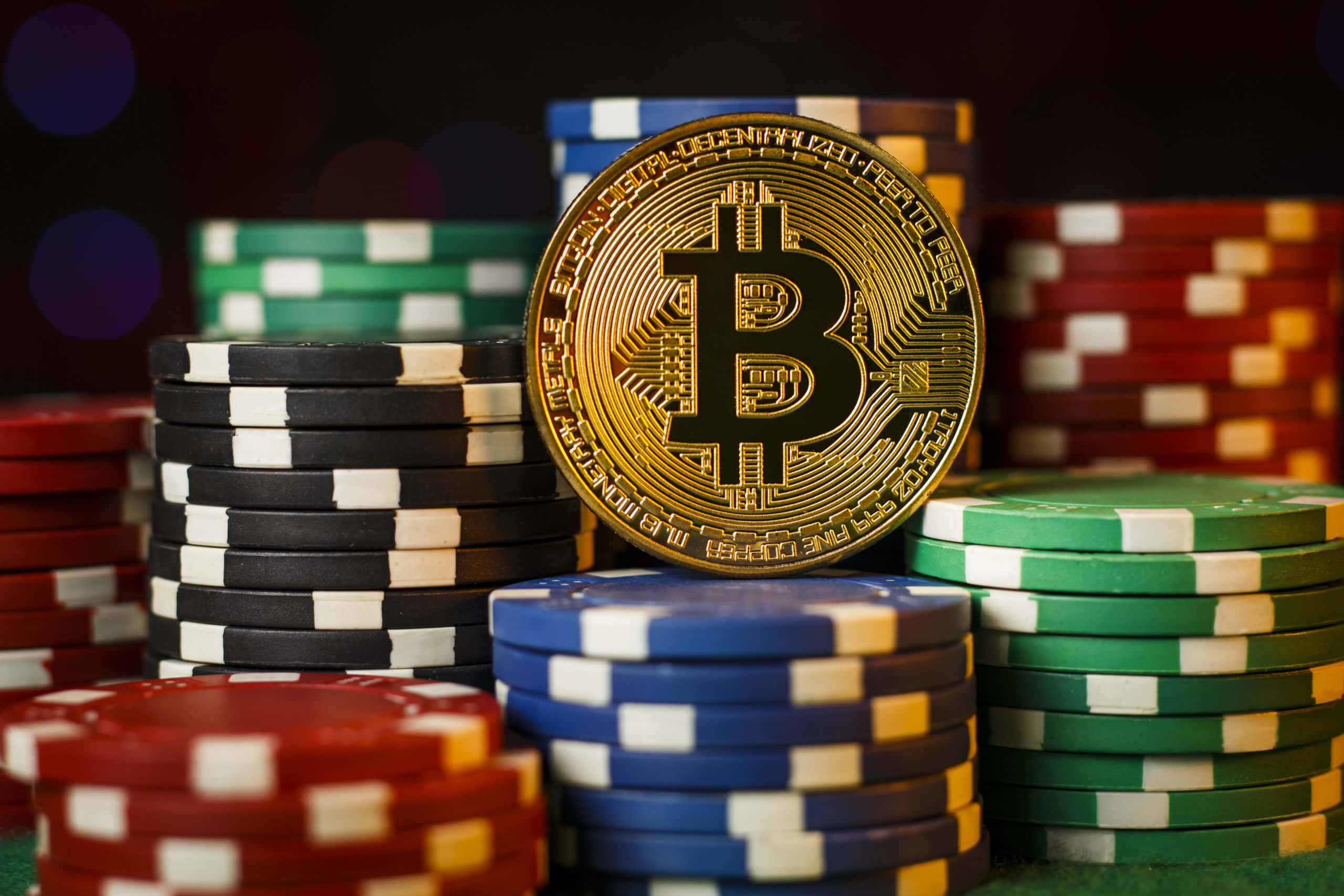比特幣 桌布 壁紙 — 一枚金色比特幣硬幣放在綠色絨布面上的彩色籌碼堆上，背後有模糊的散景燈光。