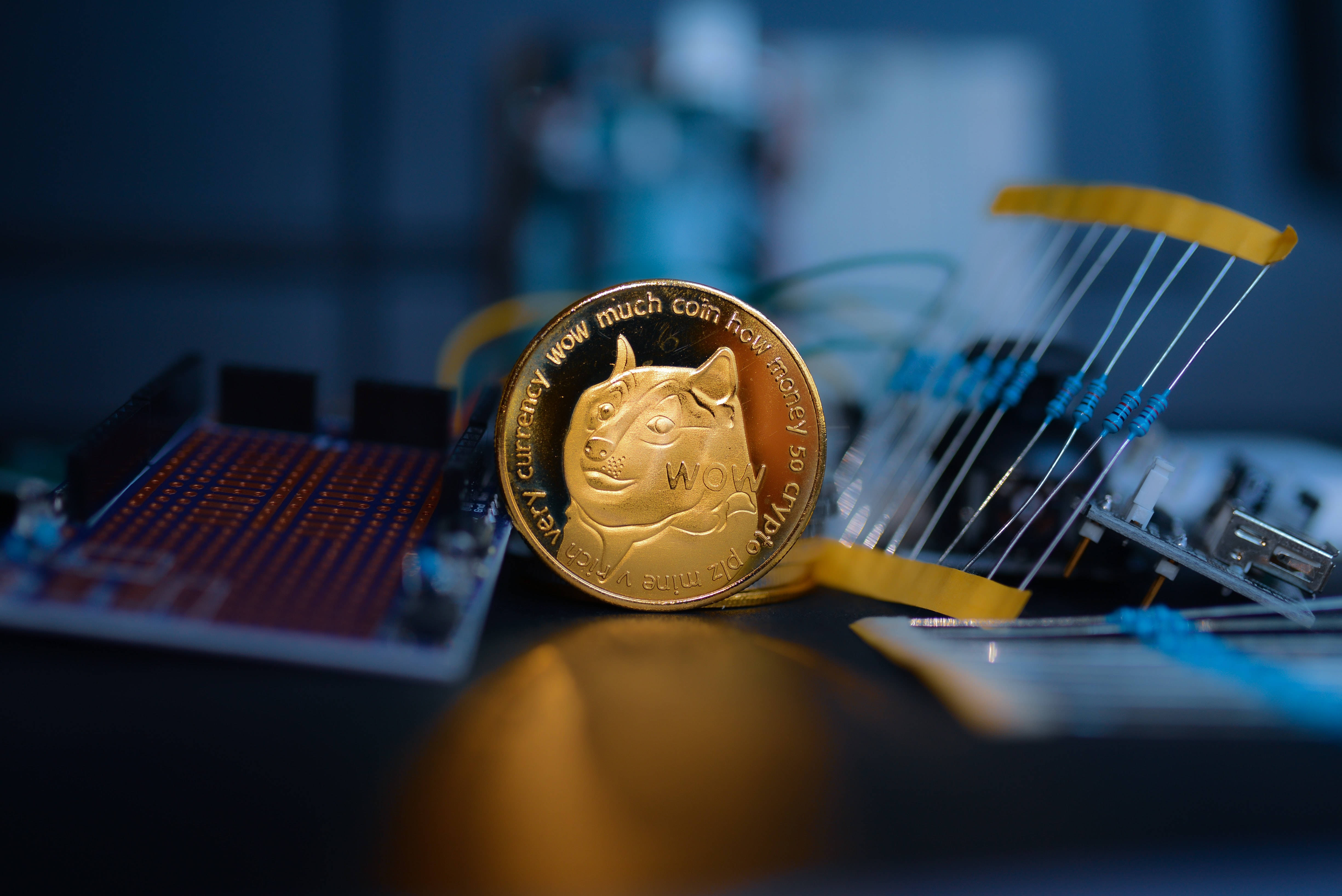 Dogecoin 桌布 壁紙 — 一枚金色狗狗幣放在帶有藍色電線和電子元件的電路板上，處於昏暗的科技環境中。