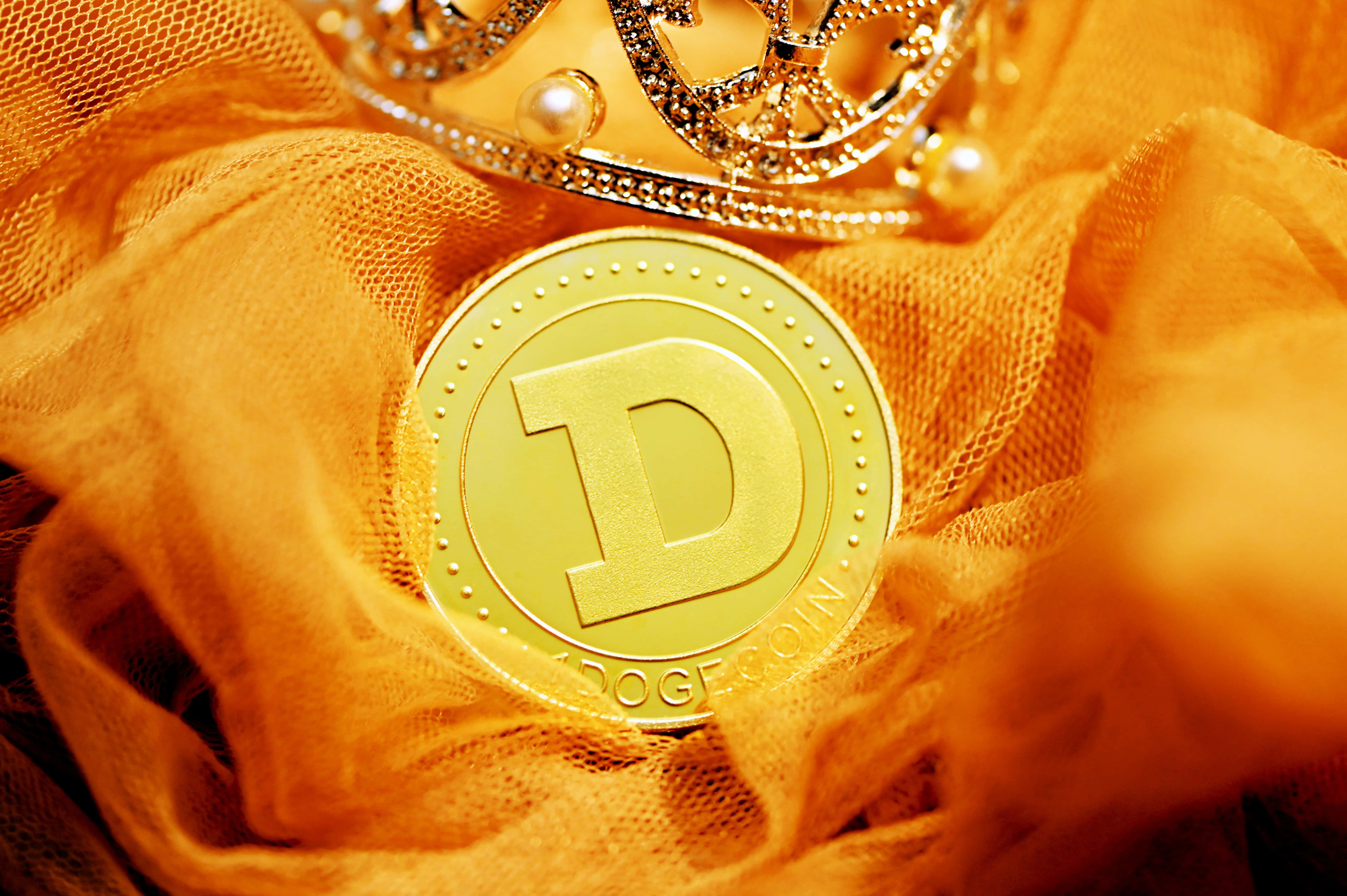 Dogecoin 桌布 壁紙 — 一枚金色狗狗幣放在橙色布料上，旁邊有一頂鑲有珠寶的皇冠，在柔和光線下溫暖地發光。