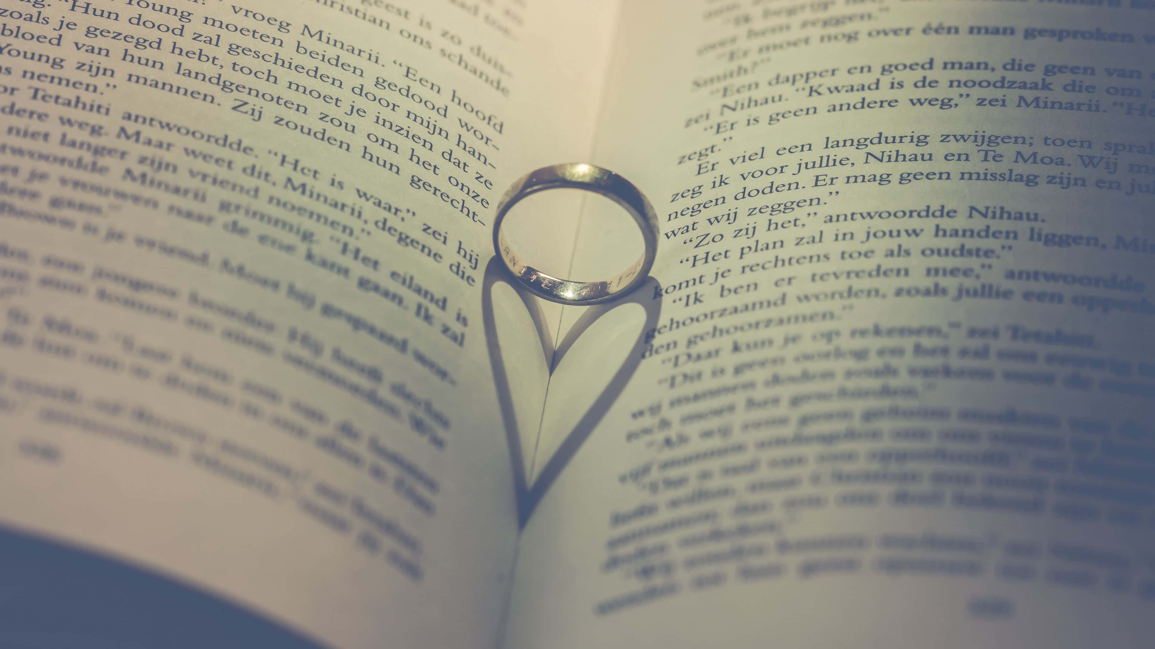 4K Love 桌布 壁紙 — 一枚銀色結婚戒指在柔和燈光下於打開的荷蘭文文字頁面上投射出心形陰影。