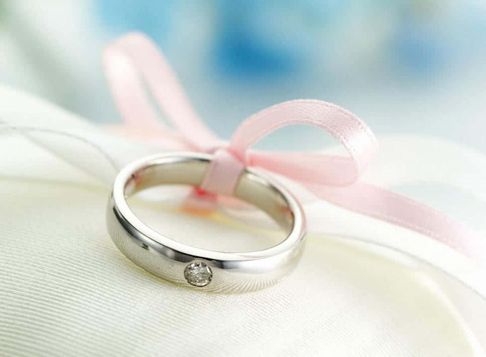 Ring 桌布 壁紙 — 一枚銀色結婚戒指鑲有一顆鑽石，放在白色布料上，繫著柔軟的粉紅色絲帶，背景是模糊的藍色。