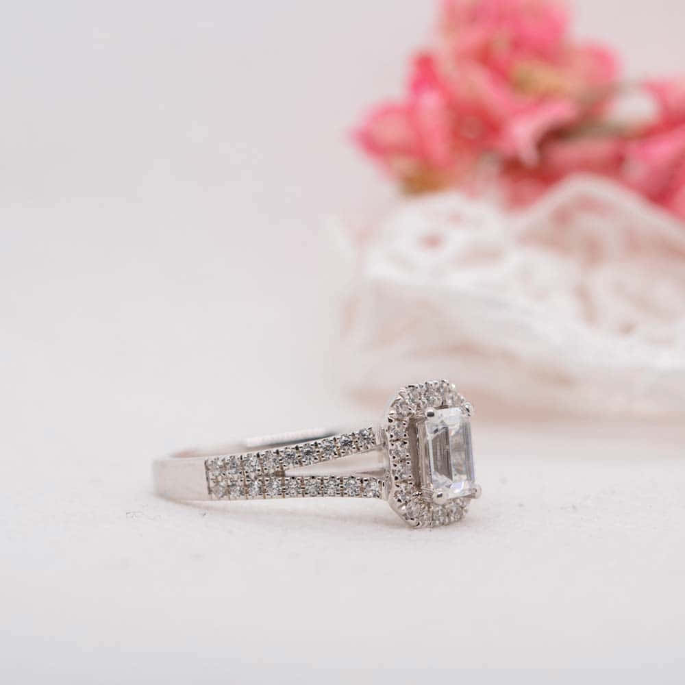 Wedding Rings 桌布 壁紙 — 一枚閃爍的祖母綠切割鑽石戒指，帶有光環和密鑲鑲嵌，放置在柔軟的白色布料上，旁邊是模糊的粉紅色花朵。