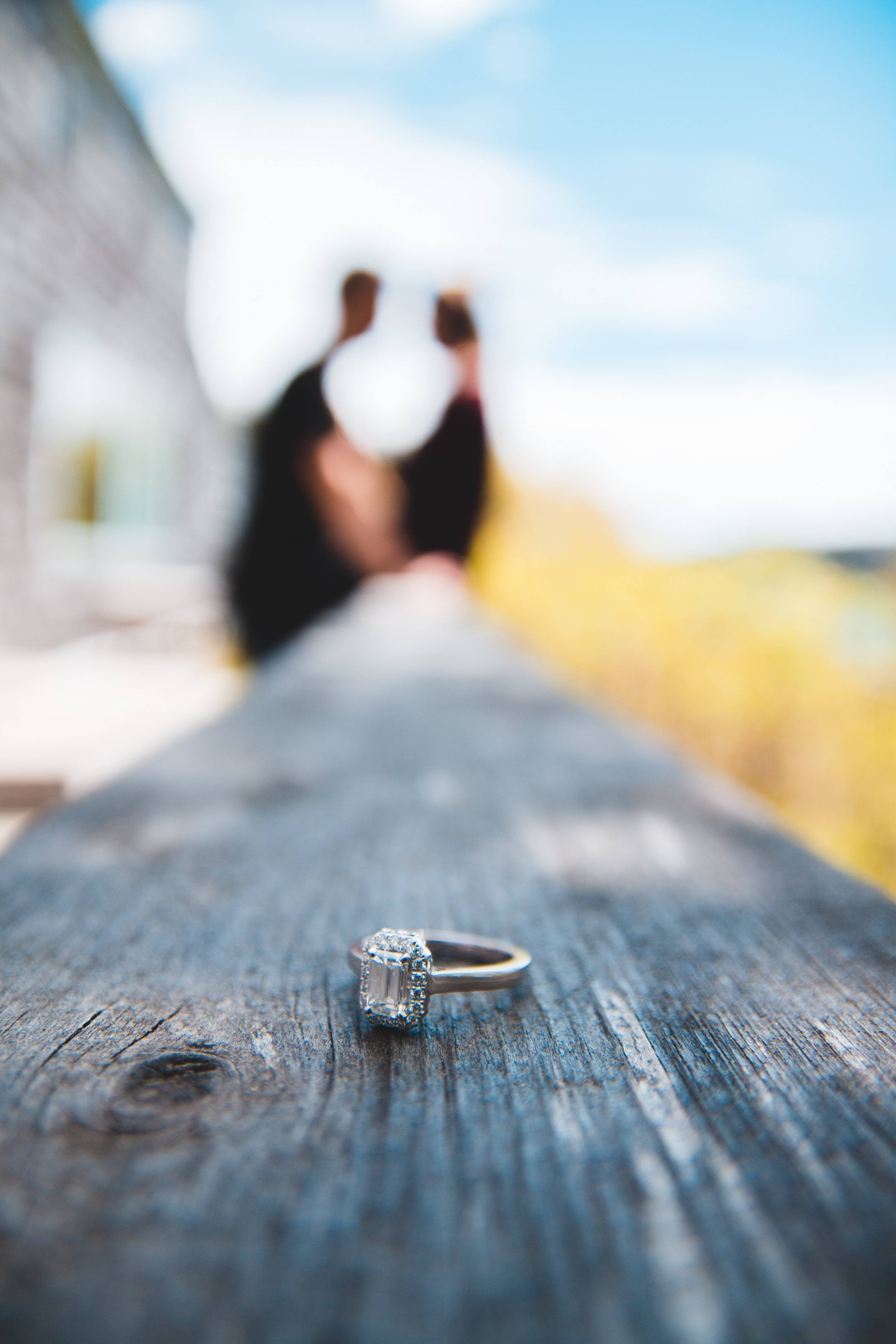 the ring 桌布 壁紙 — 一枚閃爍的祖母綠切割鑽石訂婚戒指放在風化的木頭上，背景中一對模糊的情侶在明亮的天空下擁抱。