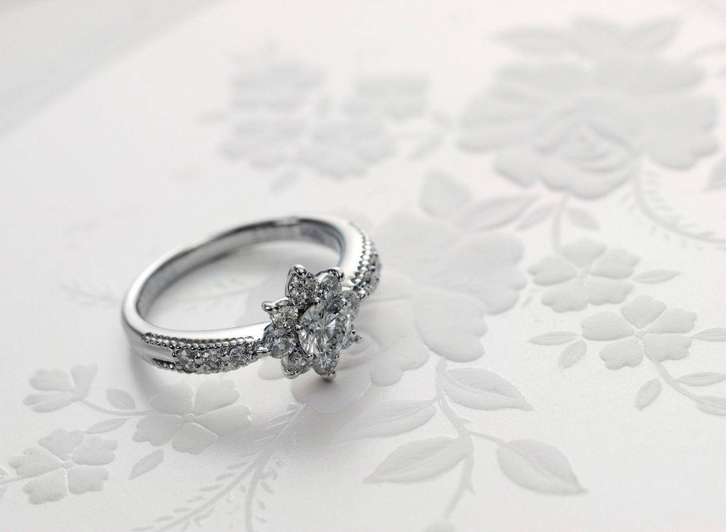 Wedding Rings 桌布 壁紙 — 一枚閃爍的鑽石訂婚戒指，帶有花卉鑽石點綴，優雅地放置在白色花卉圖案表面上。