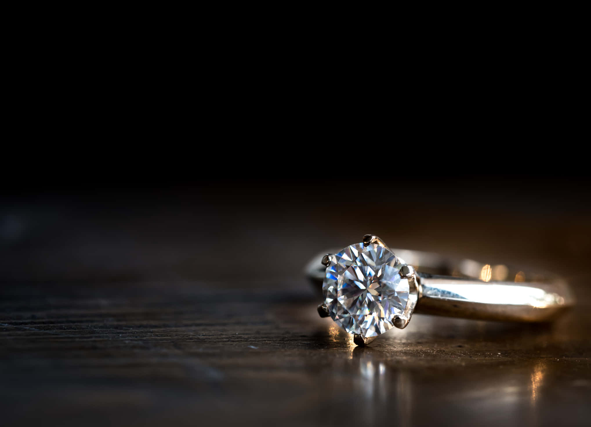 Marriage 桌布 壁紙 — 一枚閃爍的鑽石訂婚戒指在溫暖燈光下於深色木質表面上閃耀。