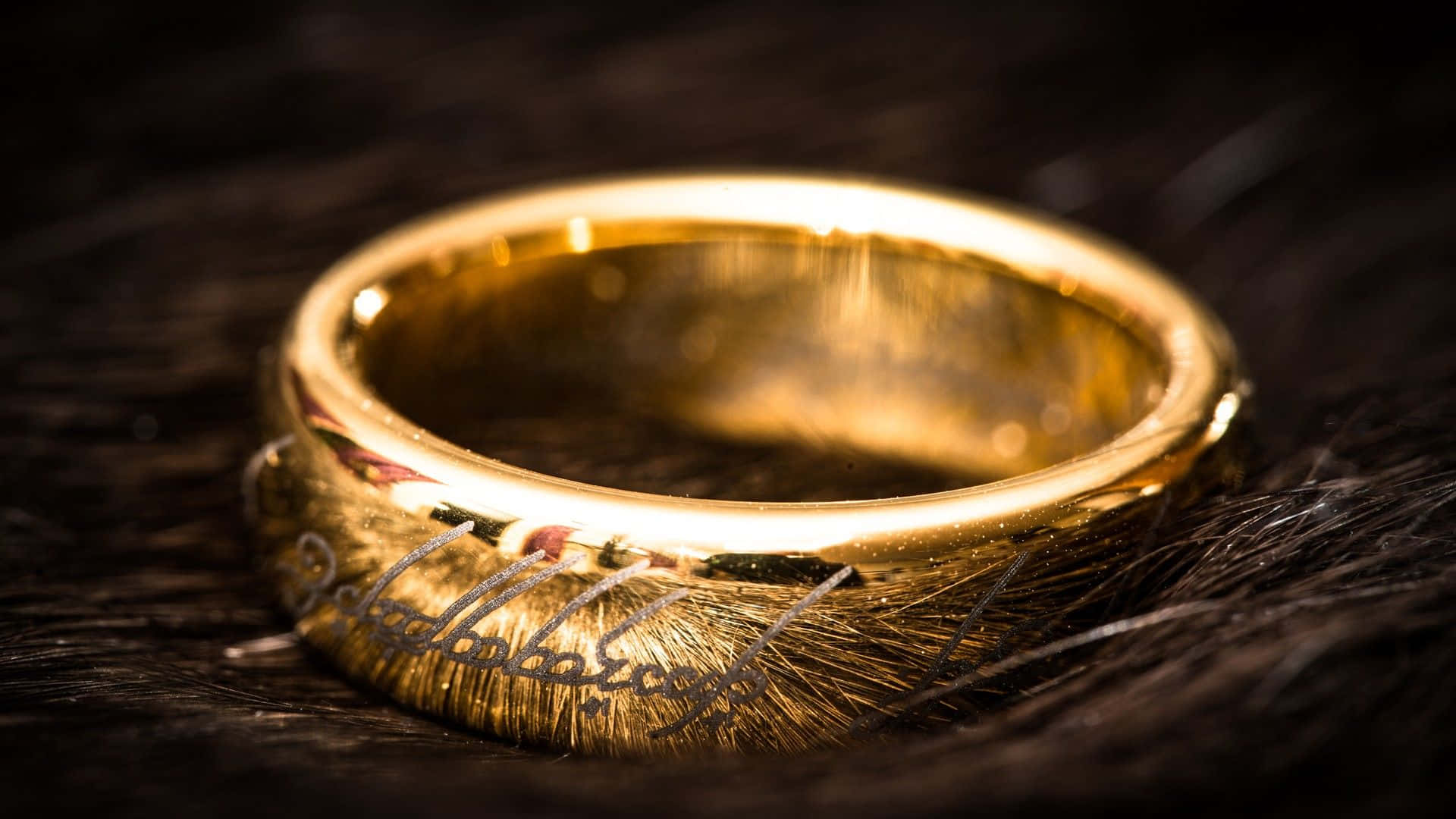 Lotr 桌布 壁紙 — 一枚閃閃發光的金戒指，上面刻有精靈文字，在溫暖聚焦的燈光下，放置在深色紋理皮毛上。