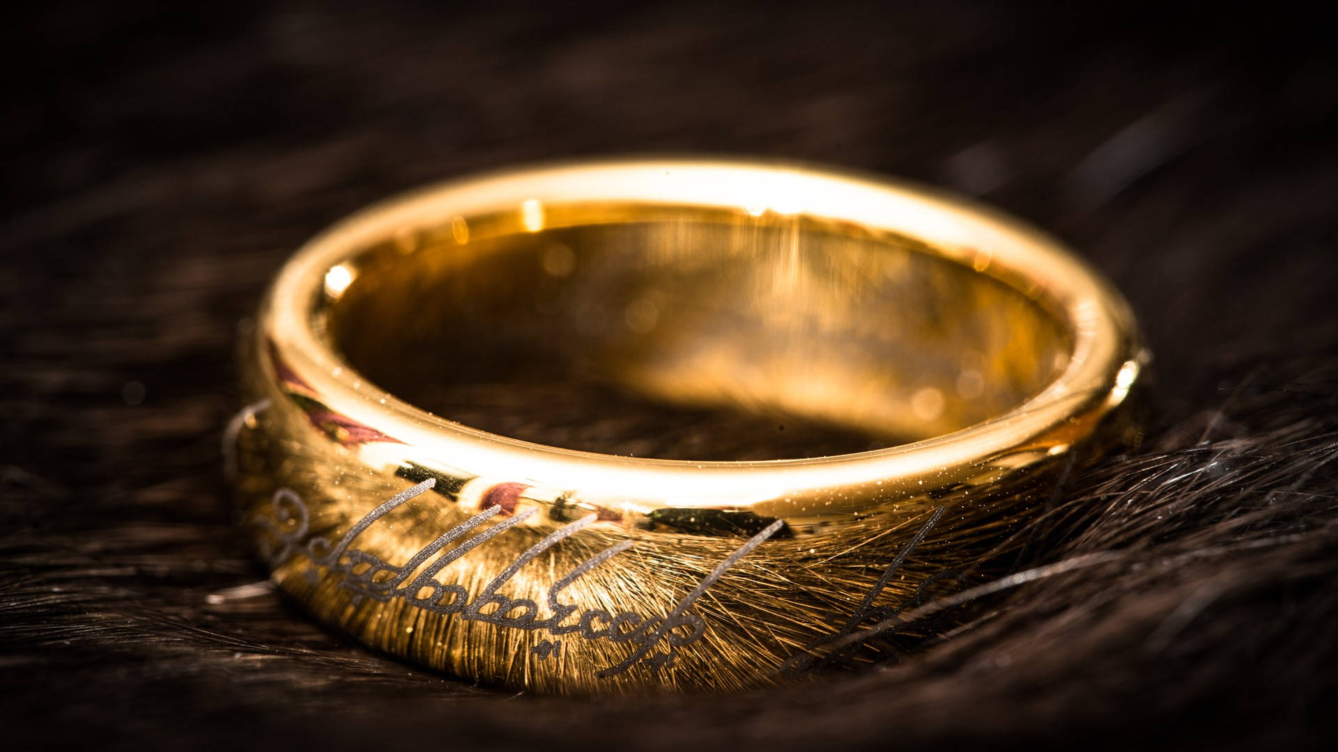 rings 桌布 壁紙 — 一枚閃閃發光的金戒指，刻有精靈文字，在溫暖的燈光下放置在深色紋理毛皮上。