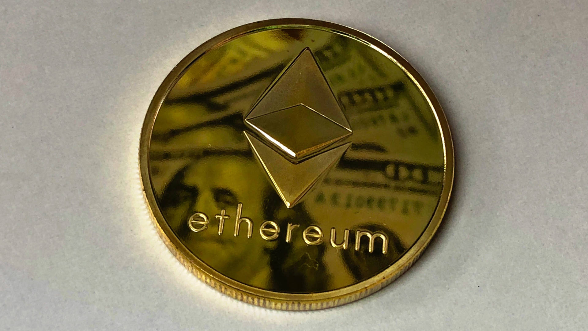 以太坊 桌布 壁紙 — 一枚閃閃發光的金色以太幣硬幣放在模糊的美鈔背景上。