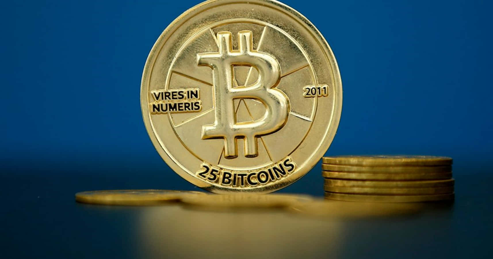 比特幣 桌布 壁紙 — 一枚閃閃發光的金色比特幣硬幣，上面刻有「25 BITCOINS」和「2011」，直立在一堆硬幣旁邊，背景是深藍色。