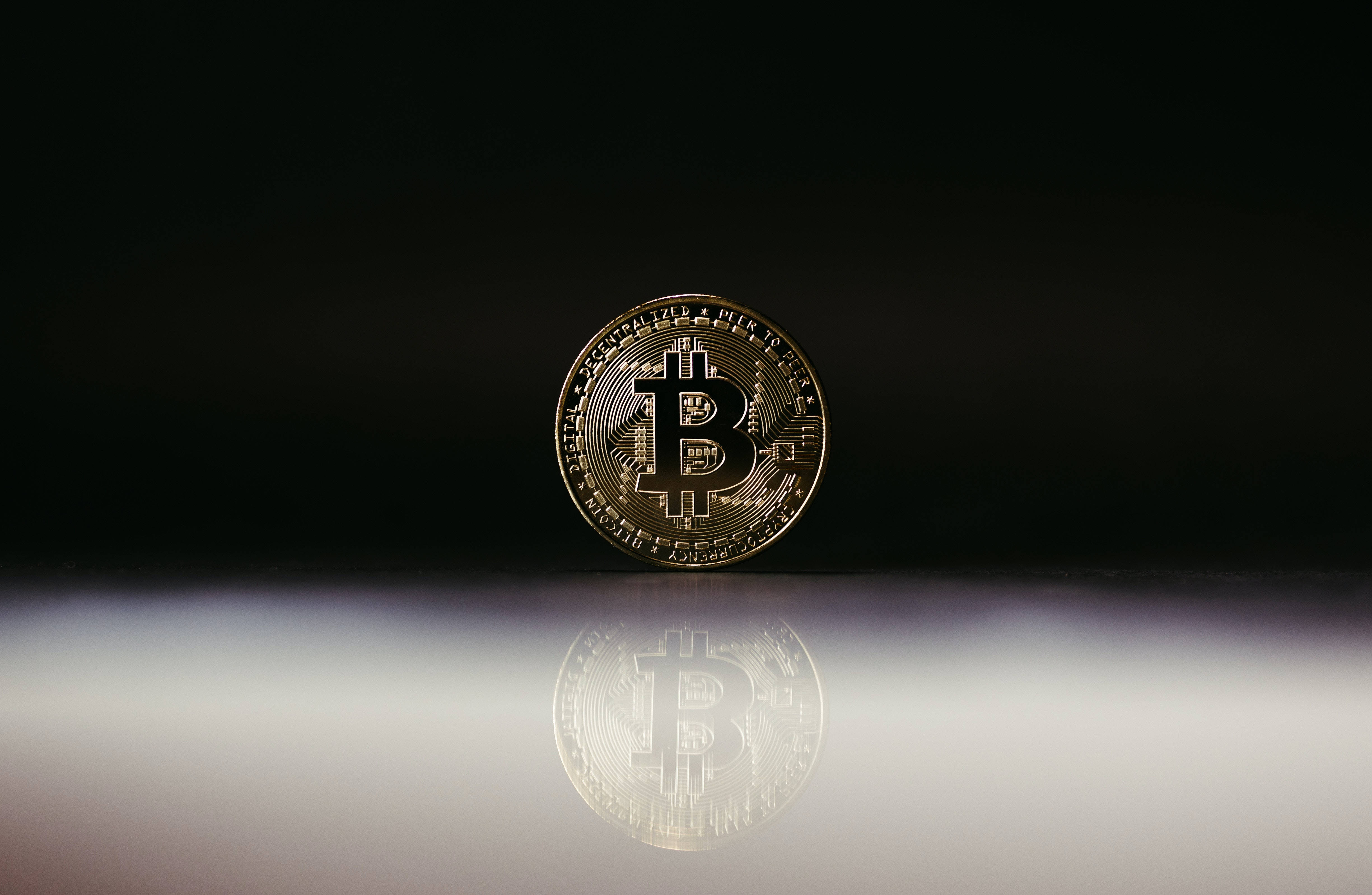 Cryptocurrency 桌布 壁紙 — 一枚閃閃發光的金色比特幣硬幣放在反光表面上，背景是純黑色。